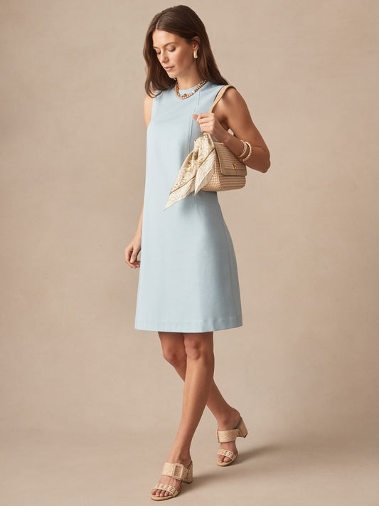 Woman wearing a sky blue Alicia Dress by J.McLaughlin, styled with beige sandals and a woven handbag with a scarf, showcasing effortlessly stylish, enduring designs and innovative fabrics.