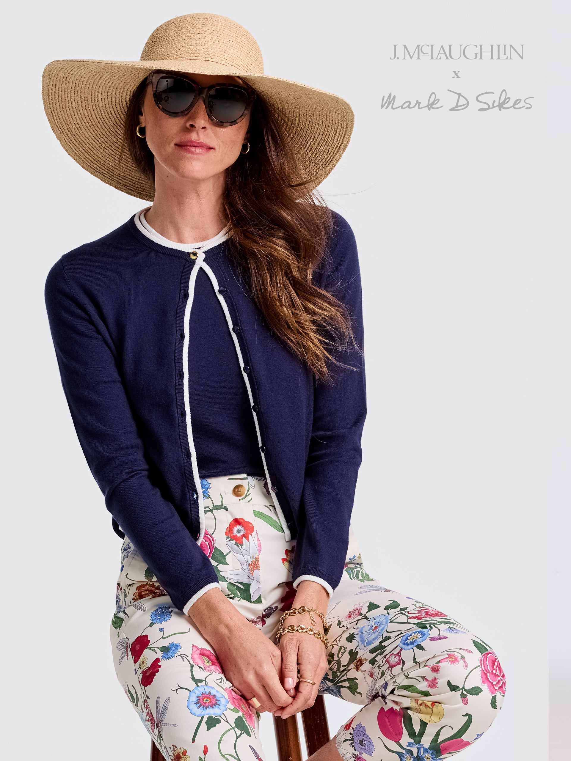 Aileen Cardigan by J.McLaughlin in navy with white trim, paired with floral pants, styled with a wide-brimmed hat and sunglasses, showcasing effortlessly stylish, enduring designs.