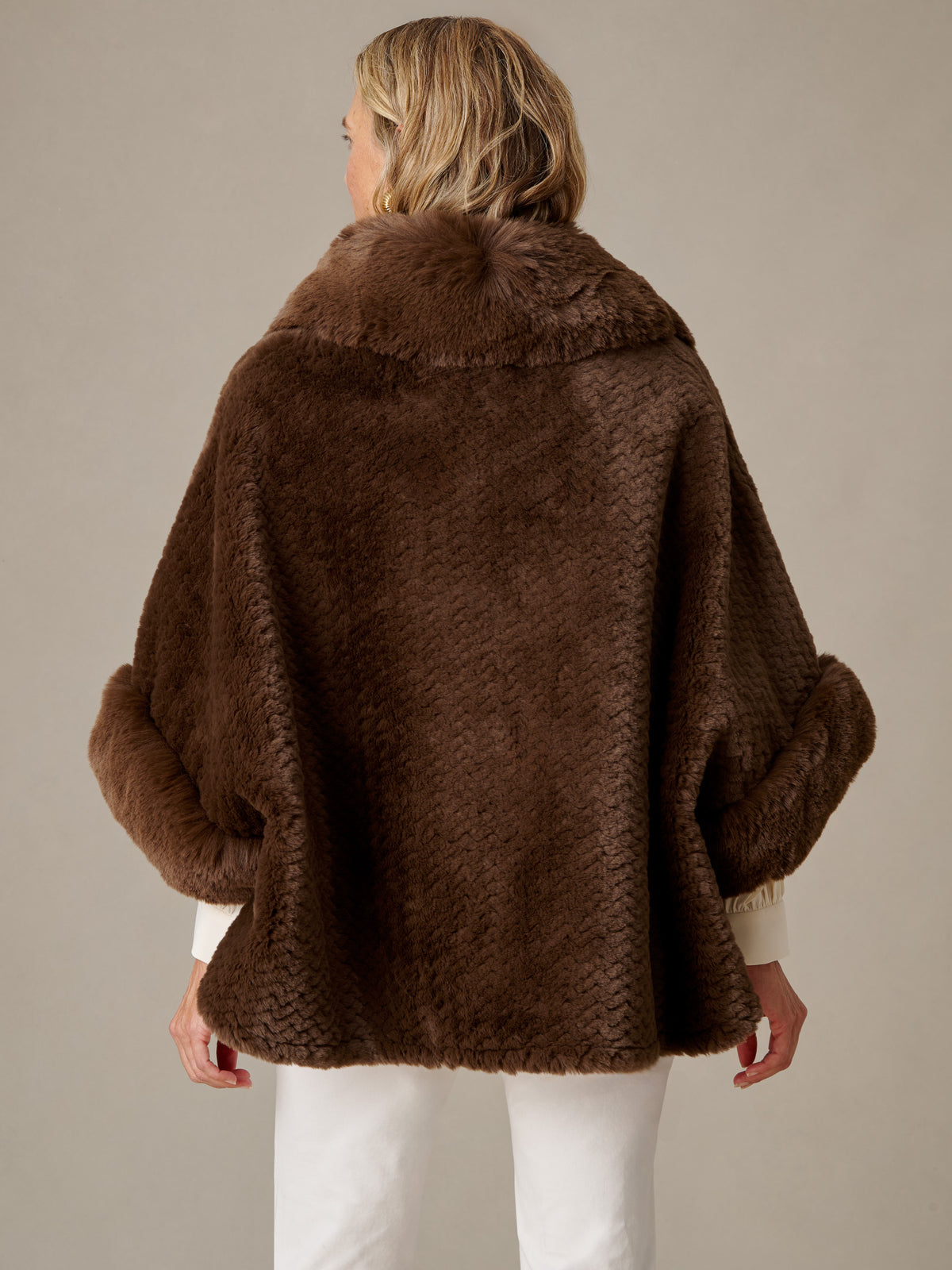 Andie Faux Fur Cape by J.McLaughlin shown from the back in rich brown faux fur with a cozy collar and wide sleeves styled over white pants