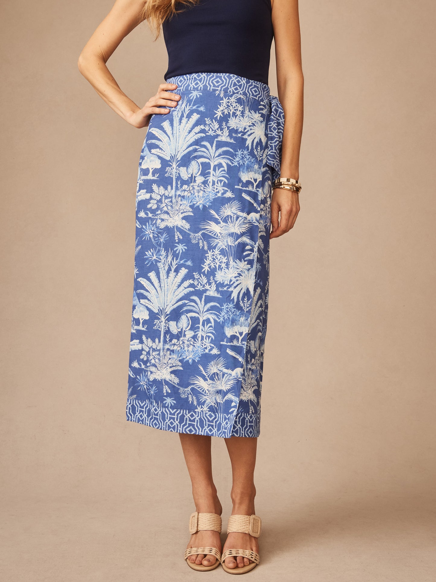A woman wearing the Ardis Linen Blend Skirt in Garden Isle by J.McLaughlin, featuring a blue and white tropical print, paired with a navy top and beige sandals, showcasing effortlessly stylish, enduring designs with innovative fabrics for a legacy of timeless fashion. A destination for defining style. Life, Well-Lived.