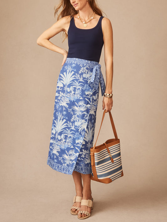 Woman wearing a navy top paired with the Ardis Linen Blend Skirt in Garden Isle by J.McLaughlin, featuring a blue and white tropical print, styled with a striped bag and sandals, showcasing effortlessly stylish, enduring designs with innovative fabrics and a legacy of timeless fashion at a destination for defining style. Life, Well-Lived.