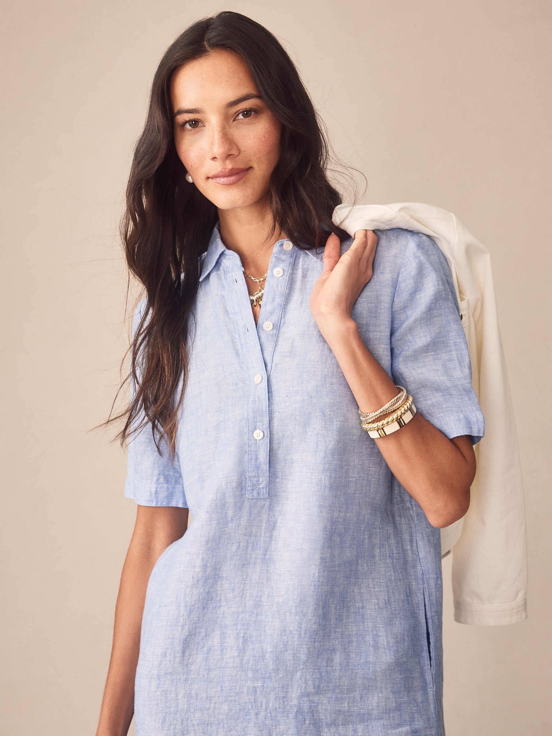 Woman wearing a light blue Arissa Linen Dress by J.McLaughlin, accessorized with gold bracelets and layered necklaces, holding a white jacket over her shoulder, showcasing effortlessly stylish, enduring designs and innovative fabrics by a destination for defining style. Life, Well-Lived.