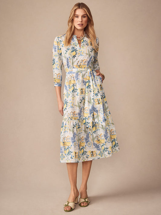 A model wearing the Cerina Dress in Amalfi Coast by J.McLaughlin, featuring effortlessly stylish, enduring designs with a light blue and yellow scenic print, embodying a legacy of timeless fashion. A destination for defining style. Life, Well-Lived.