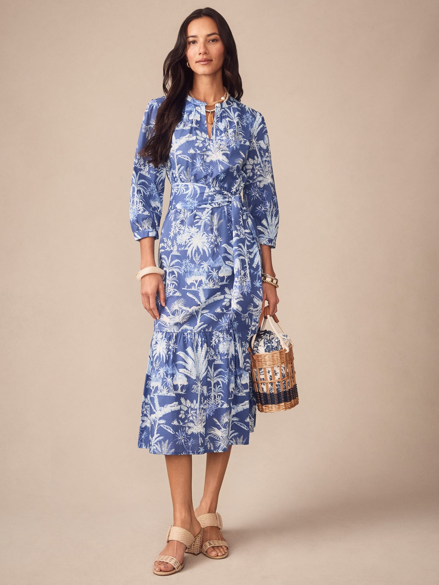 A model wears the Cerina Dress in Garden Isle by J.McLaughlin, featuring an effortlessly stylish blue tropical print dress with a belted waist, complemented by natural woven sandals and a wicker bag, embodying enduring designs and innovative fabrics. A destination for defining style. Life, Well-Lived.