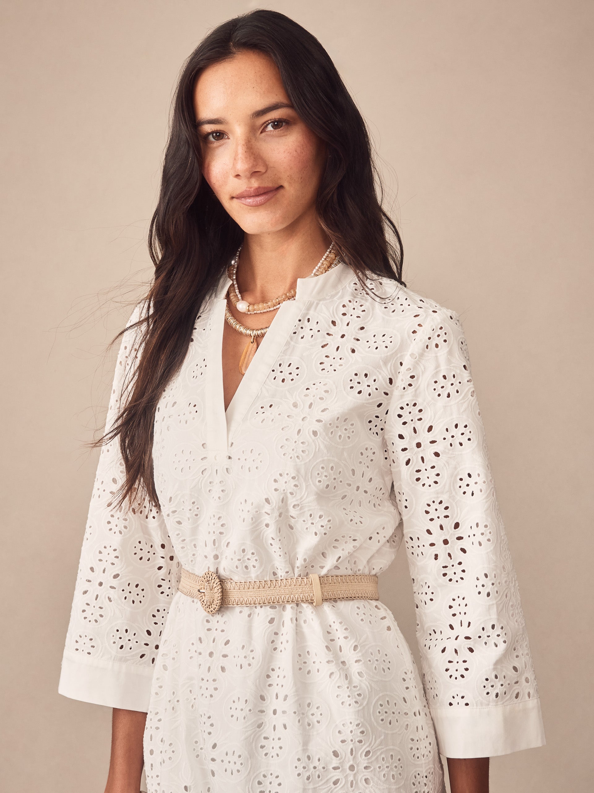 A woman wearing a white Cindy Eyelet Dress by J.McLaughlin featuring intricate eyelet detailing, a beige woven belt, and layered pearl necklaces, embodying effortlessly stylish and enduring designs.