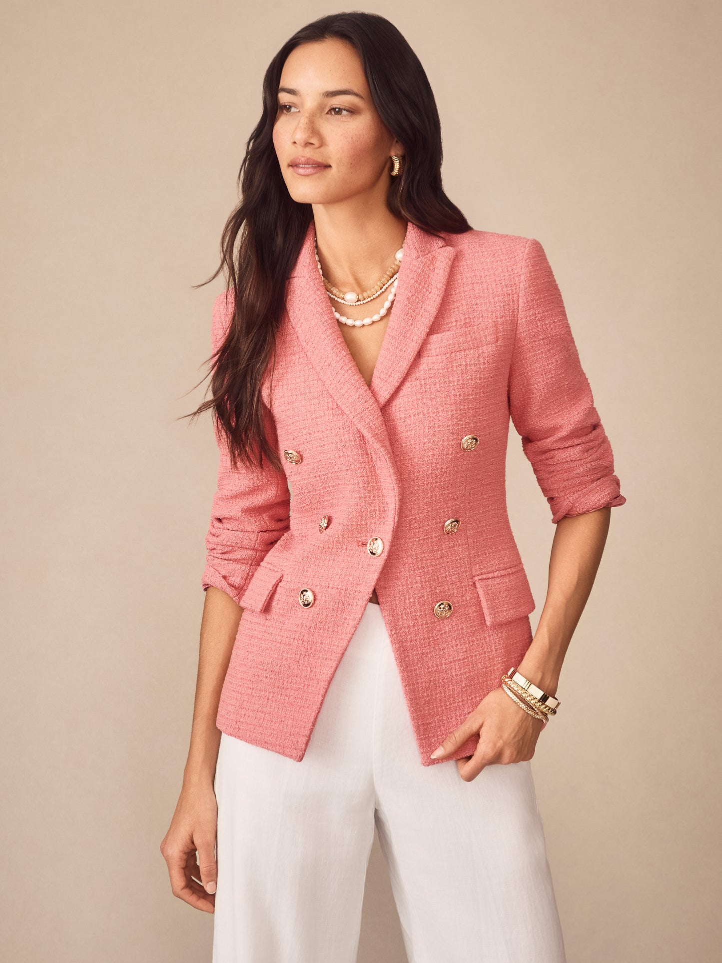 Woman wearing a pink Genevieve Cotton Bouclé Blazer by J.McLaughlin with gold buttons, styled with white pants and layered pearl necklaces, showcasing effortlessly stylish enduring designs.