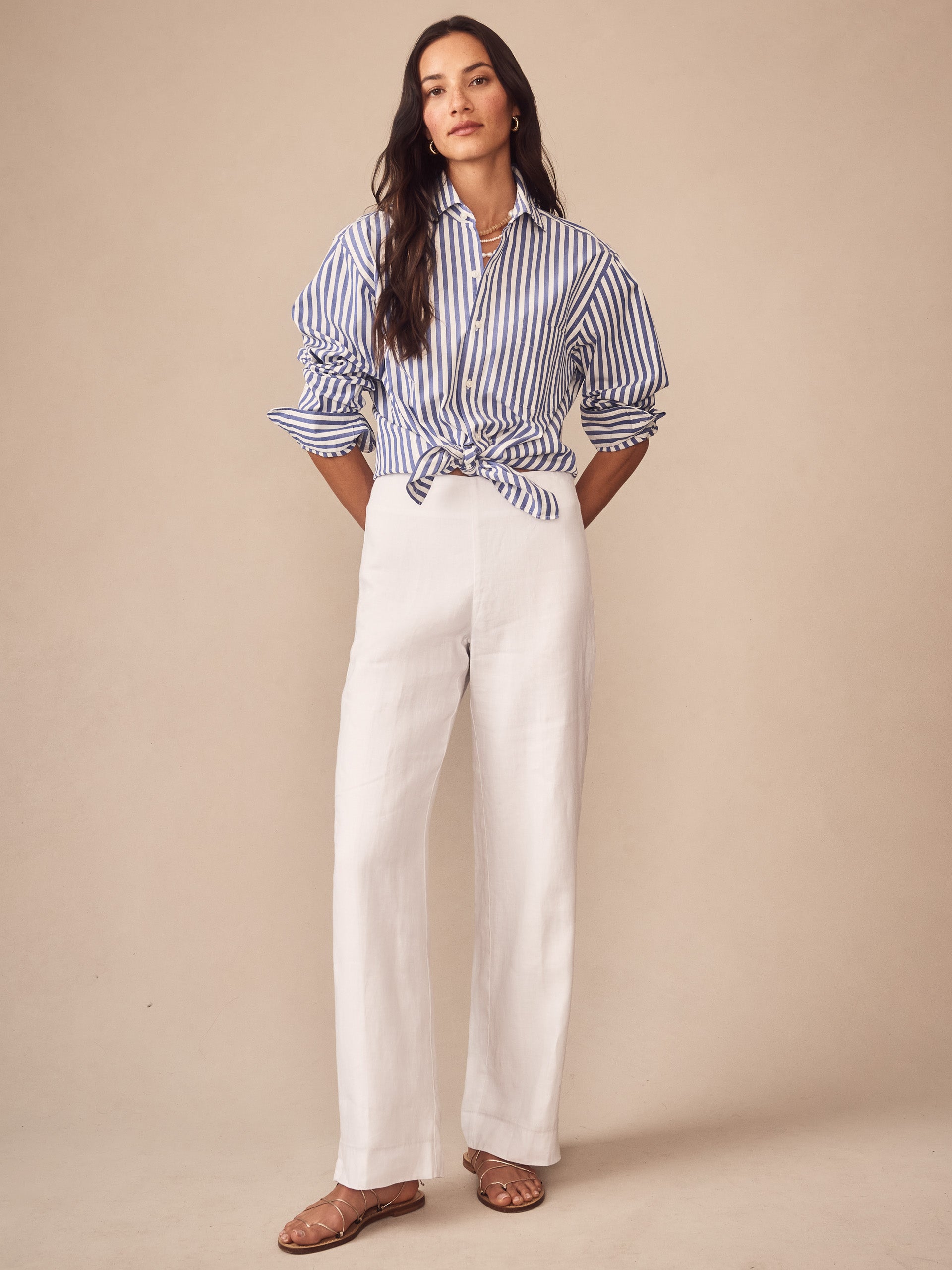 Woman wearing Carter Linen Pants in white with a blue and white striped shirt tied at the waist and brown sandals, showcasing J.McLaughlin’s effortlessly stylish, enduring designs and innovative fabrics.