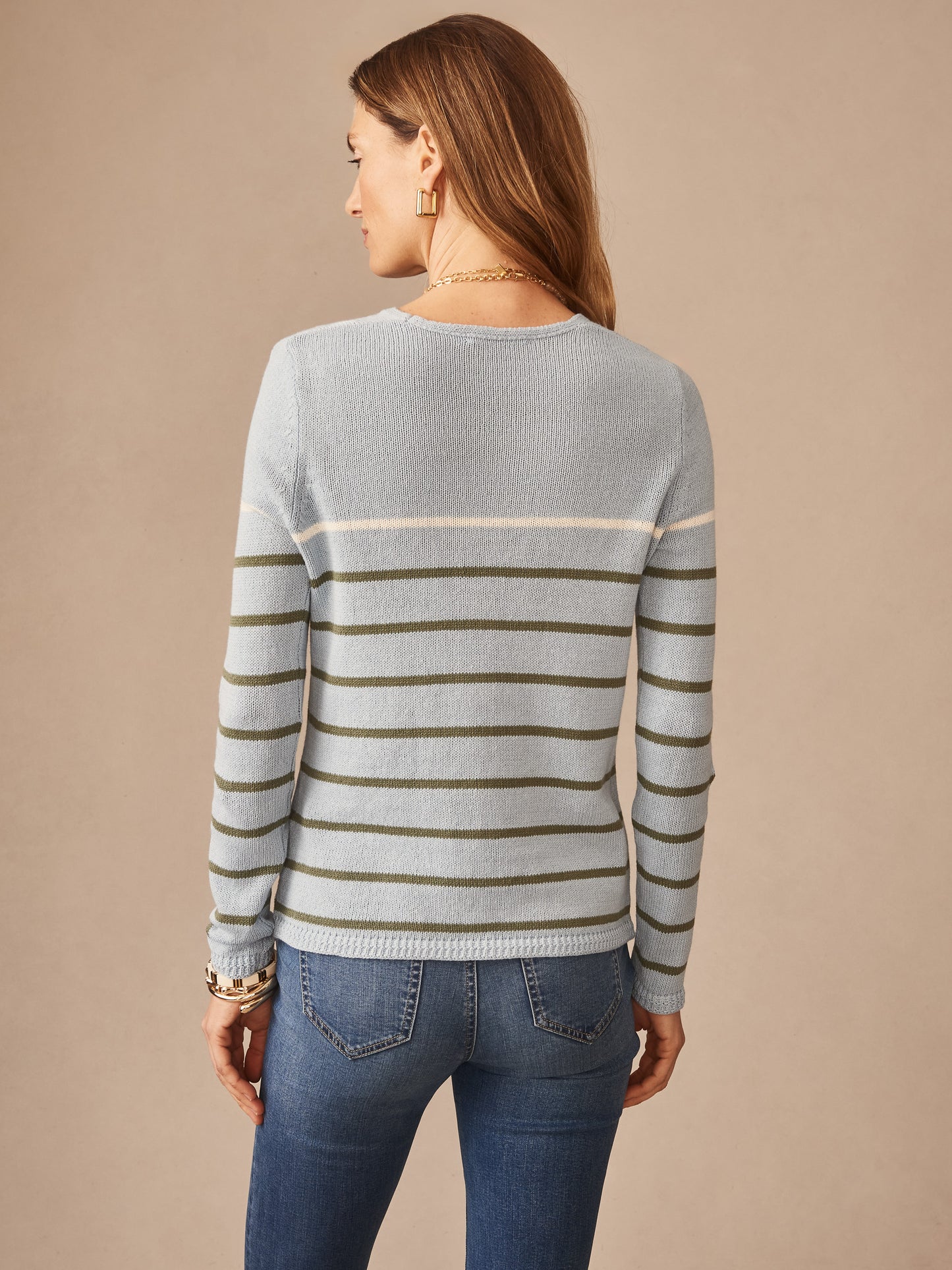 Woman wearing a Ramona Linen Blend Sweater in Stripe by J.McLaughlin, featuring olive and cream horizontal stripes on a light blue background, paired with jeans. Effortlessly stylish with enduring designs and innovative fabrics, embodying a legacy of timeless fashion. A destination for defining style. Life, Well-Lived.