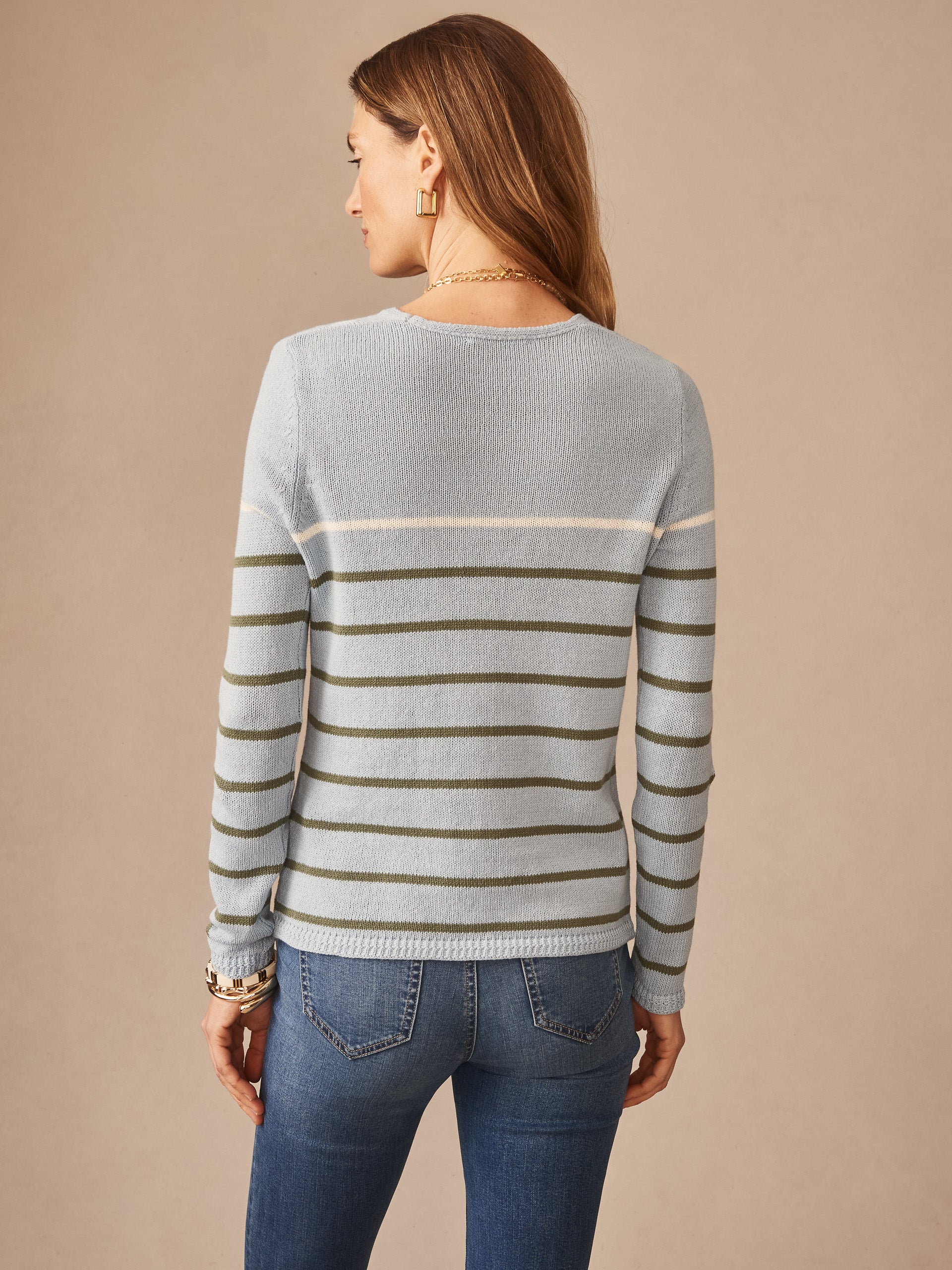 Woman wearing a Ramona Linen Blend Sweater in Stripe by J.McLaughlin, featuring olive and cream horizontal stripes on a light blue background, paired with jeans. Effortlessly stylish with enduring designs and innovative fabrics, embodying a legacy of timeless fashion. A destination for defining style. Life, Well-Lived.