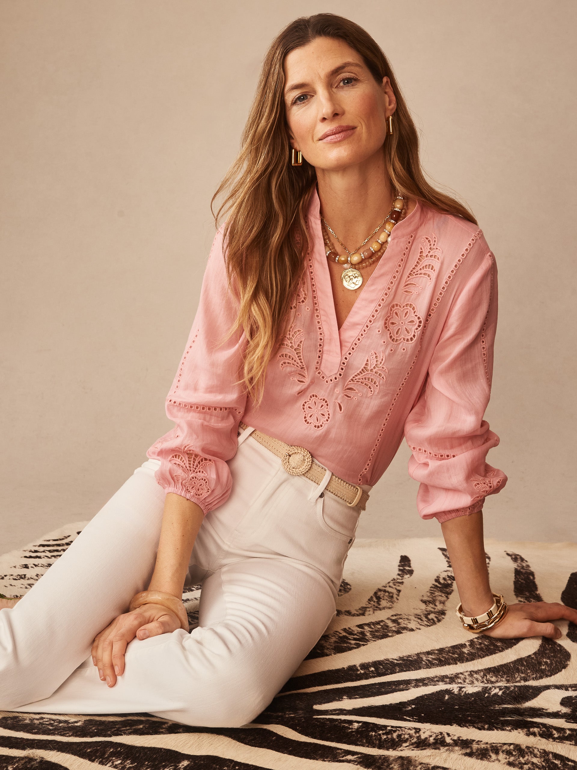 Woman wearing a pink Varsha Blouse by J.McLaughlin featuring delicate embroidery and lace details, paired with white pants and gold accessories, showcasing effortlessly stylish, enduring designs and innovative fabrics. A destination for defining style. Life, Well-Lived.