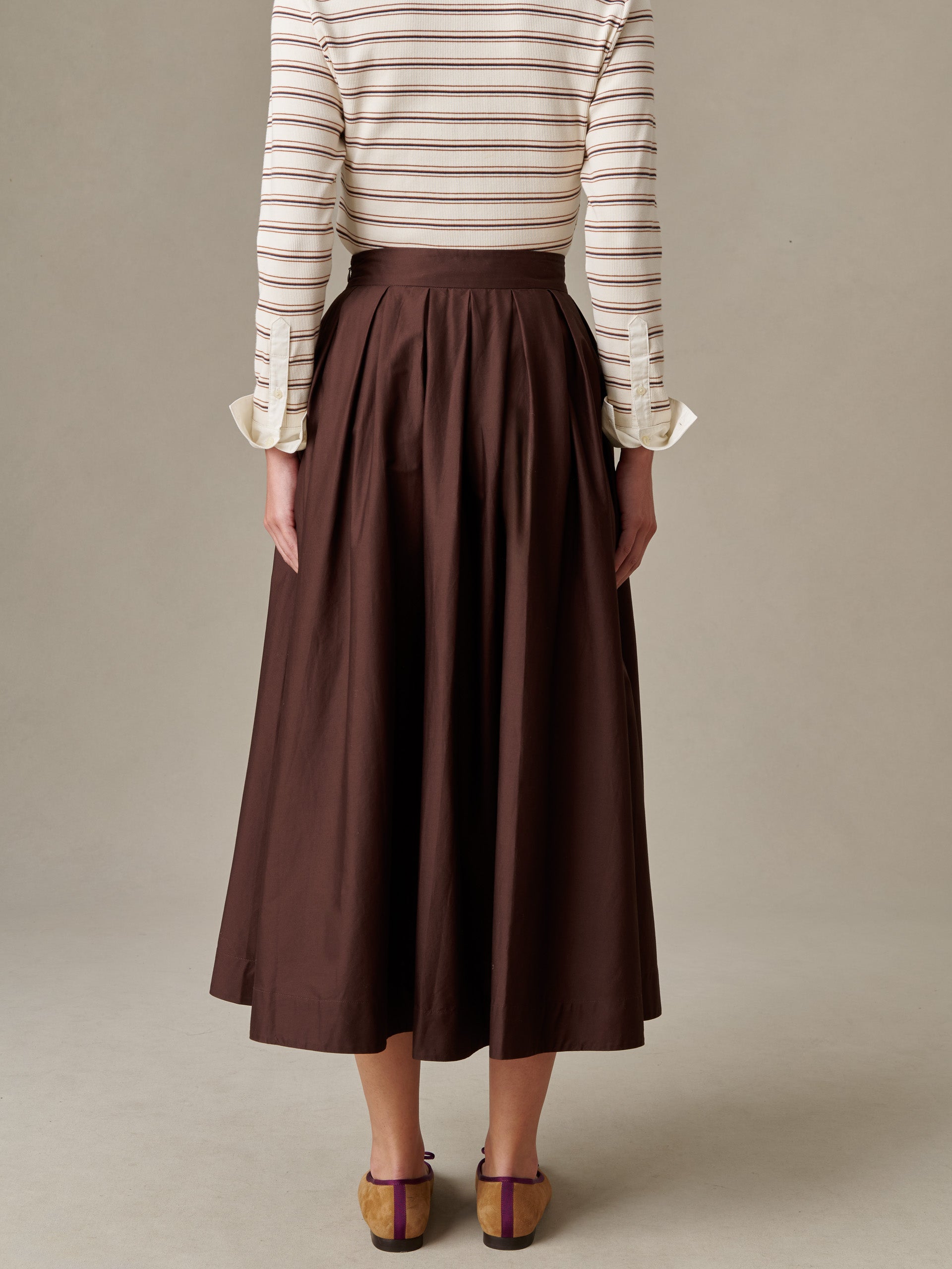 Back view of a model wearing the Arabella Skirt by J.McLaughlin, featuring a high-waisted, pleated silhouette in rich brown, styled with a striped top and heeled shoes.
