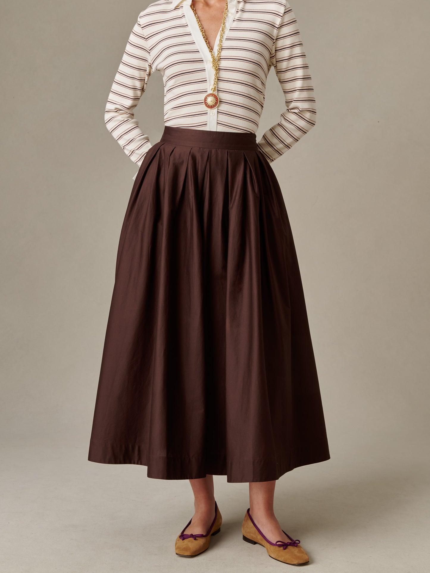 Woman wearing a high-waisted, pleated brown Arabella Skirt by J.McLaughlin paired with a striped, long-sleeve blouse and tan flats, showcasing timeless style and elegance.