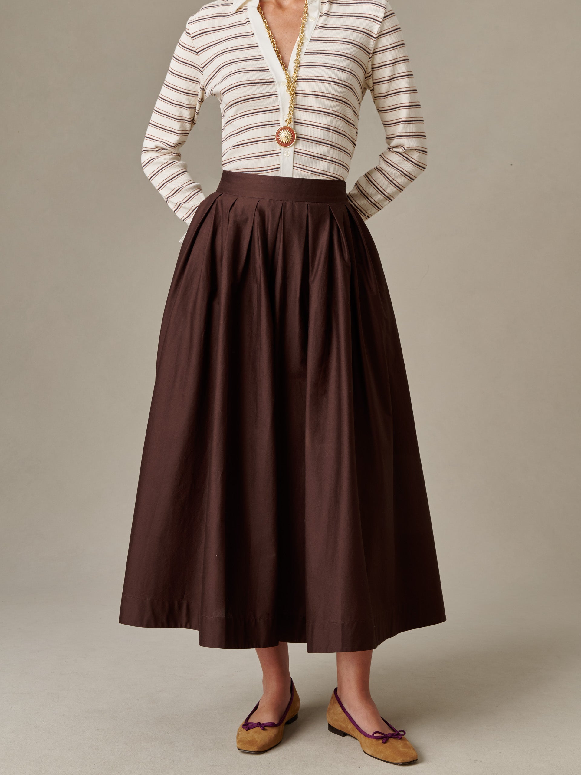 Woman wearing a high-waisted, pleated brown Arabella Skirt by J.McLaughlin paired with a striped, long-sleeve blouse and tan flats, showcasing timeless style and elegance.