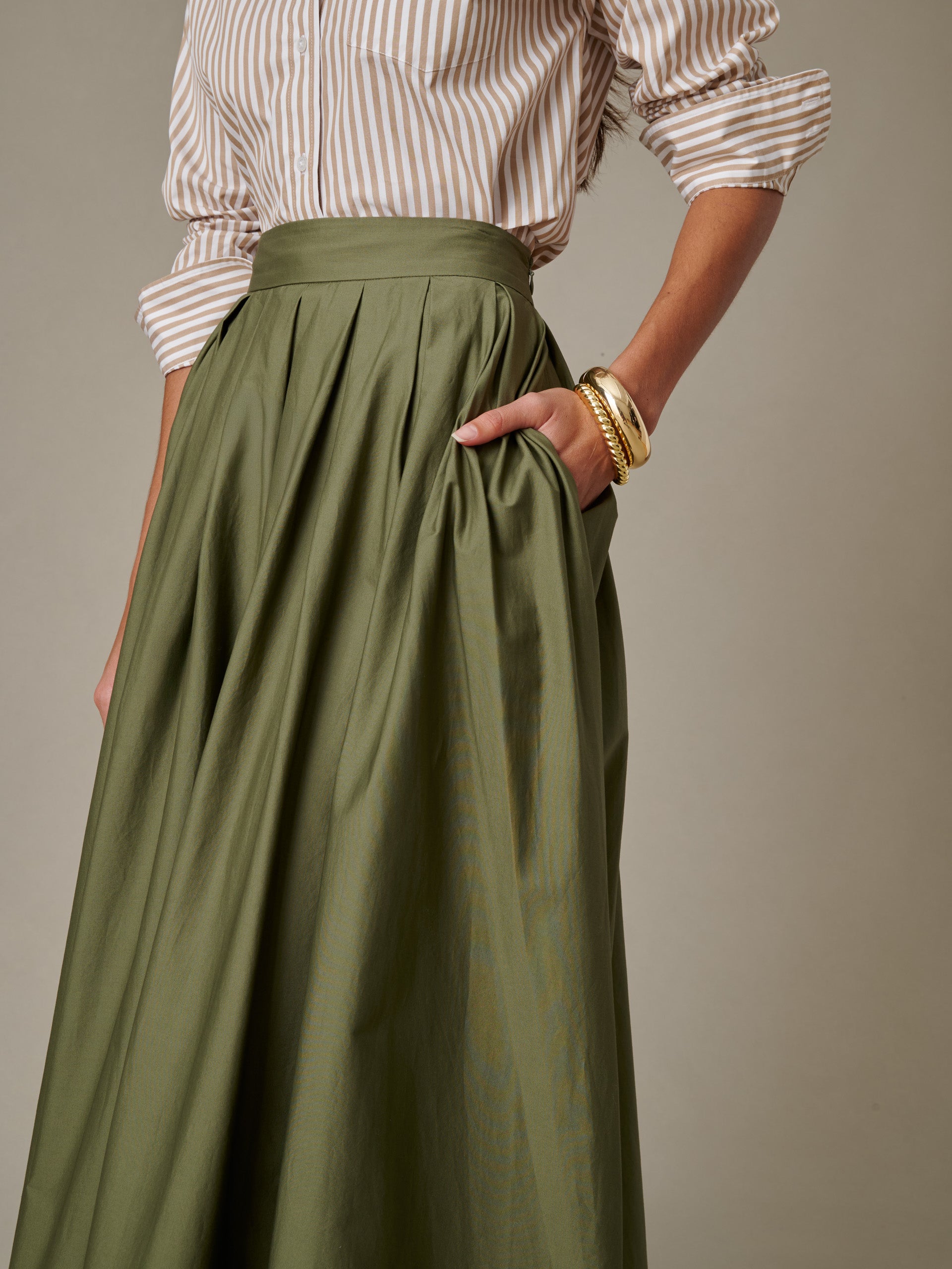 Woman wearing an olive green Arabella Skirt by J.McLaughlin paired with a striped shirt and gold bangle bracelets, showcasing timeless style and impeccable craftsmanship.