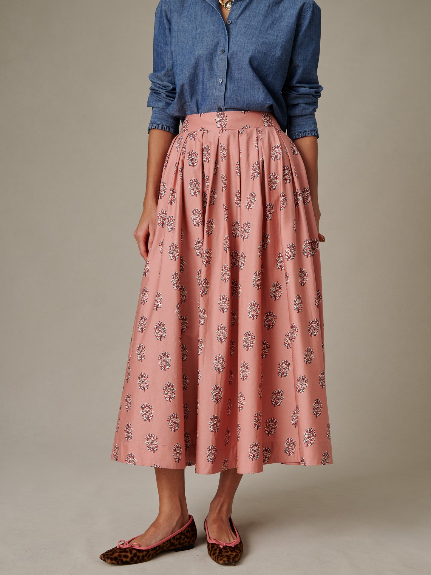 Woman wearing a pink Arabella Skirt in Heritage Bloom Cross with delicate floral pattern paired with a rolled-up sleeve chambray shirt and leopard print ballet flats by J.McLaughlin, showcasing effortlessly stylish, enduring design.