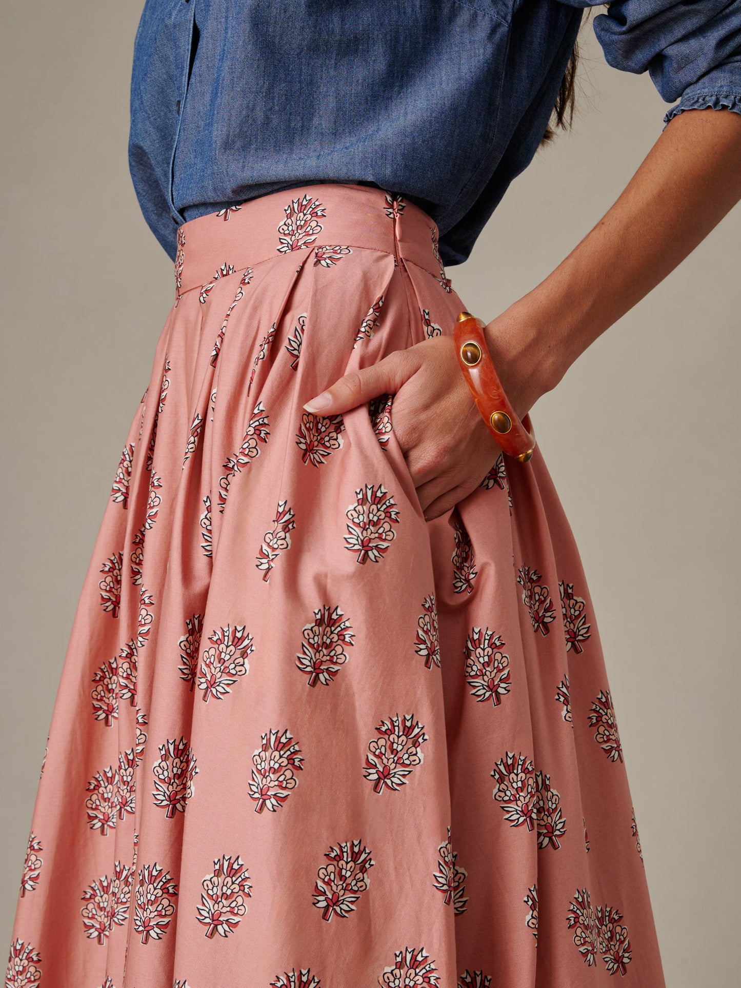 Close-up of a woman wearing the Arabella Skirt in Heritage Bloom Cross by J.McLaughlin, featuring a soft pink floral pattern paired with a blue chambray top and orange bracelet, showcasing effortlessly stylish and enduring design.