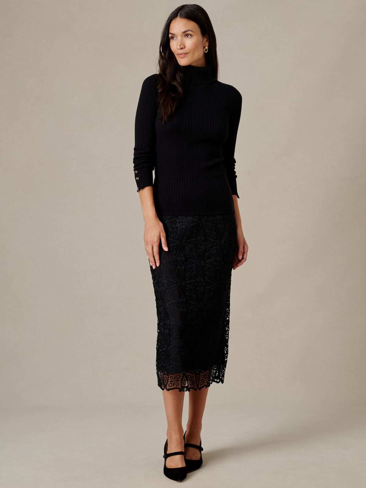 A woman models the Arlette Turtleneck by J.McLaughlin in black ribbed cotton modal with button cuffs styled with a lace midi skirt and heels