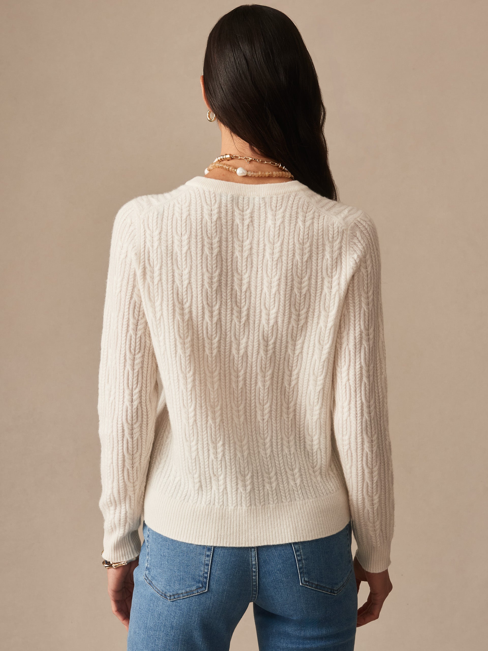 Woman wearing an ivory Ashby Cashmere Sweater by J.McLaughlin, featuring a classic cable knit design, paired with blue jeans, embodying effortlessly stylish and enduring designs in luxurious fabric.