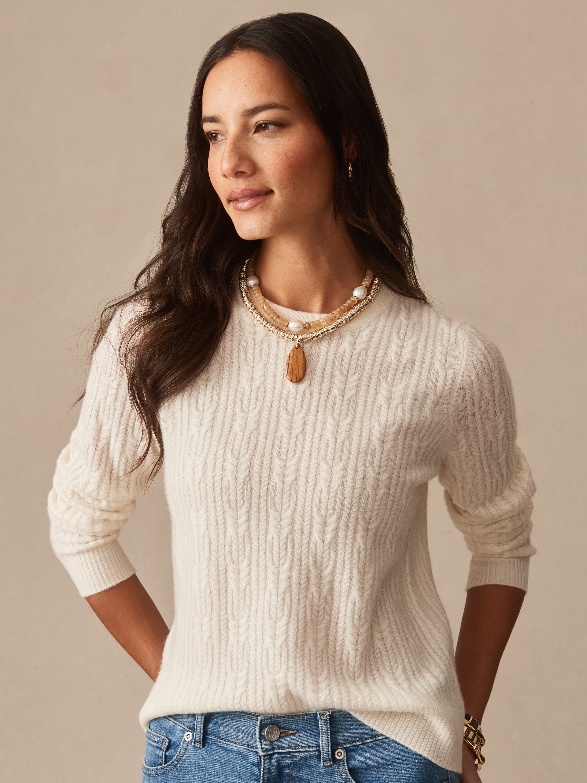 Woman wearing an ivory Ashby Cashmere Sweater by J.McLaughlin, featuring a classic cable knit design, paired with layered necklaces and casual blue jeans, embodying effortlessly stylish, enduring designs.