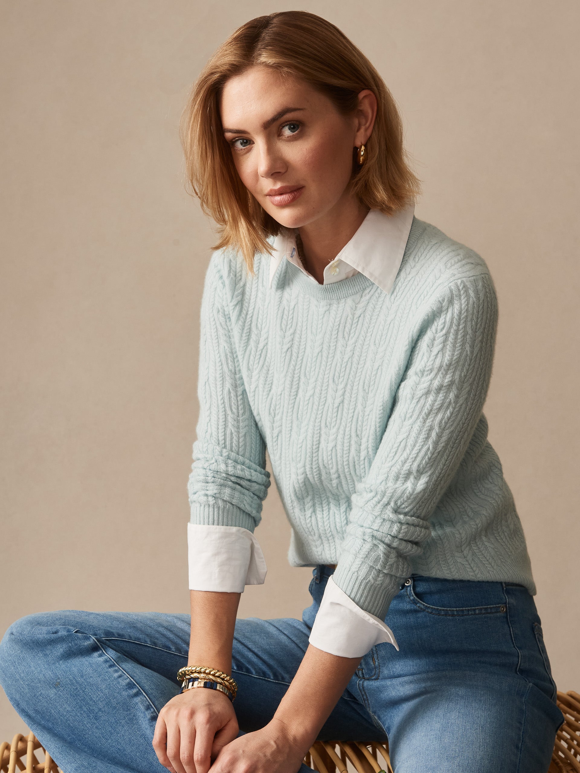 Woman wearing an effortlessly stylish Ashby Cashmere Sweater in soft aqua by J.McLaughlin, paired with a crisp white shirt and jeans, showcasing enduring designs and a legacy of timeless fashion.