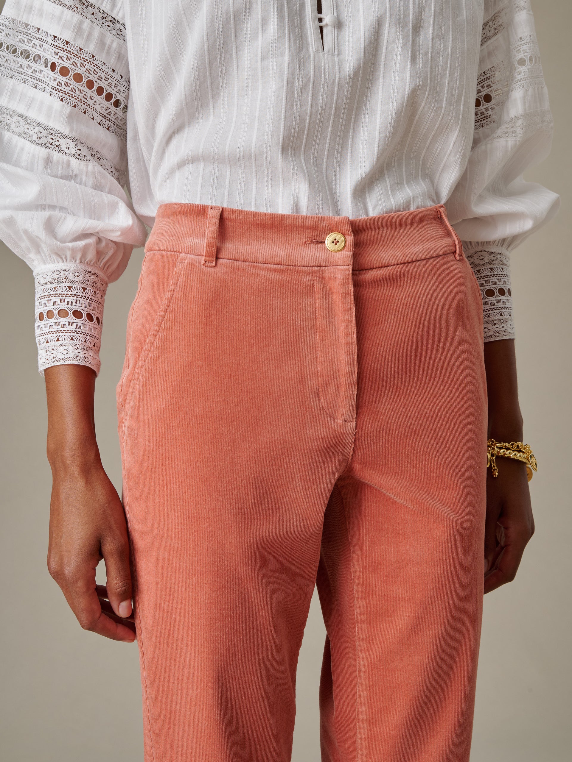 Close-up of a person wearing Avery Corduroy Pants by J.McLaughlin in a warm terracotta shade paired with a white blouse featuring lace and striped detailing, showcasing timeless style and craftsmanship.