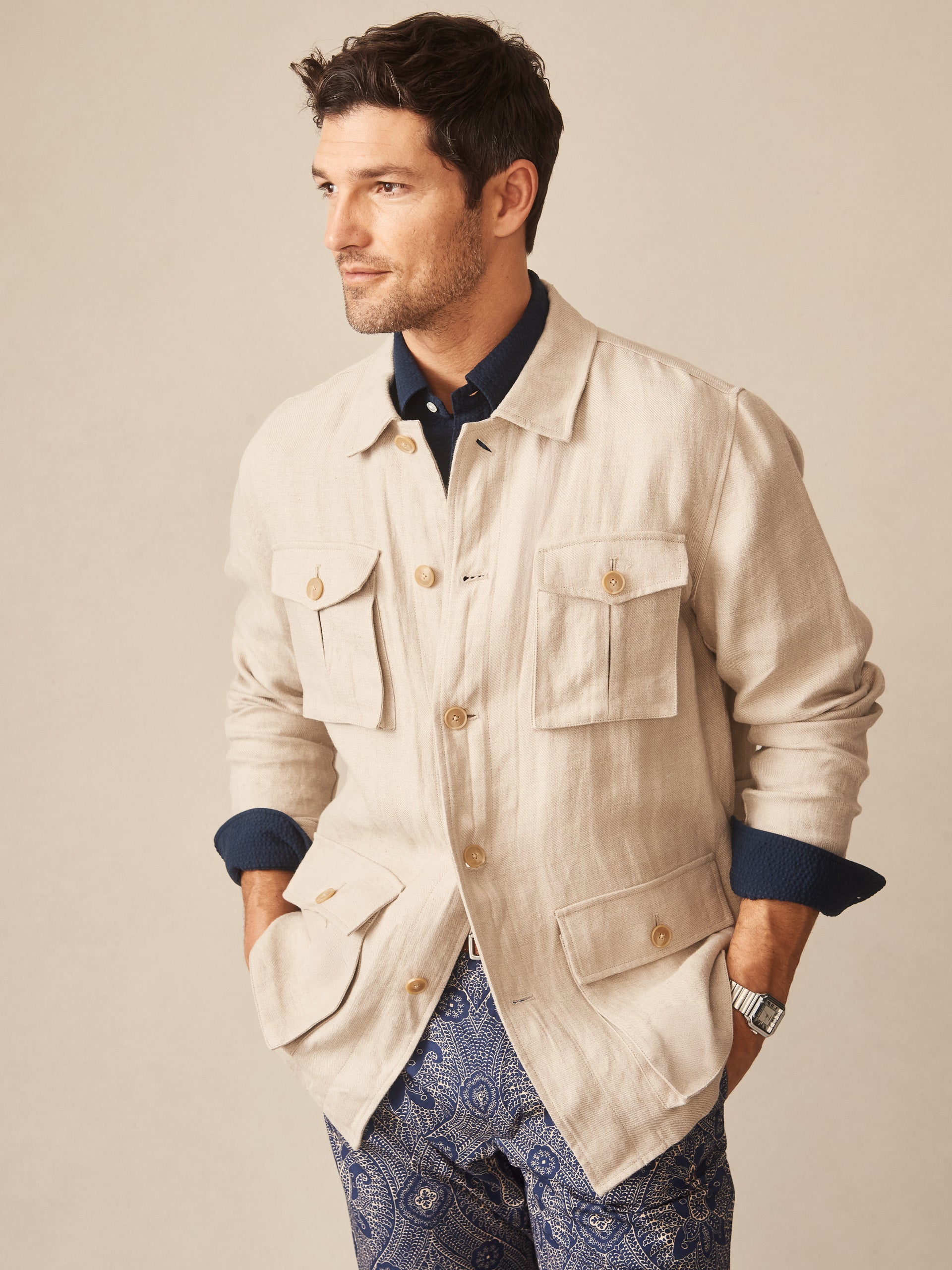 Man wearing a J.McLaughlin Aston Linen Safari Jacket in Herringbone over a navy shirt and patterned pants, showcasing effortlessly stylish, enduring designs with innovative fabrics.
