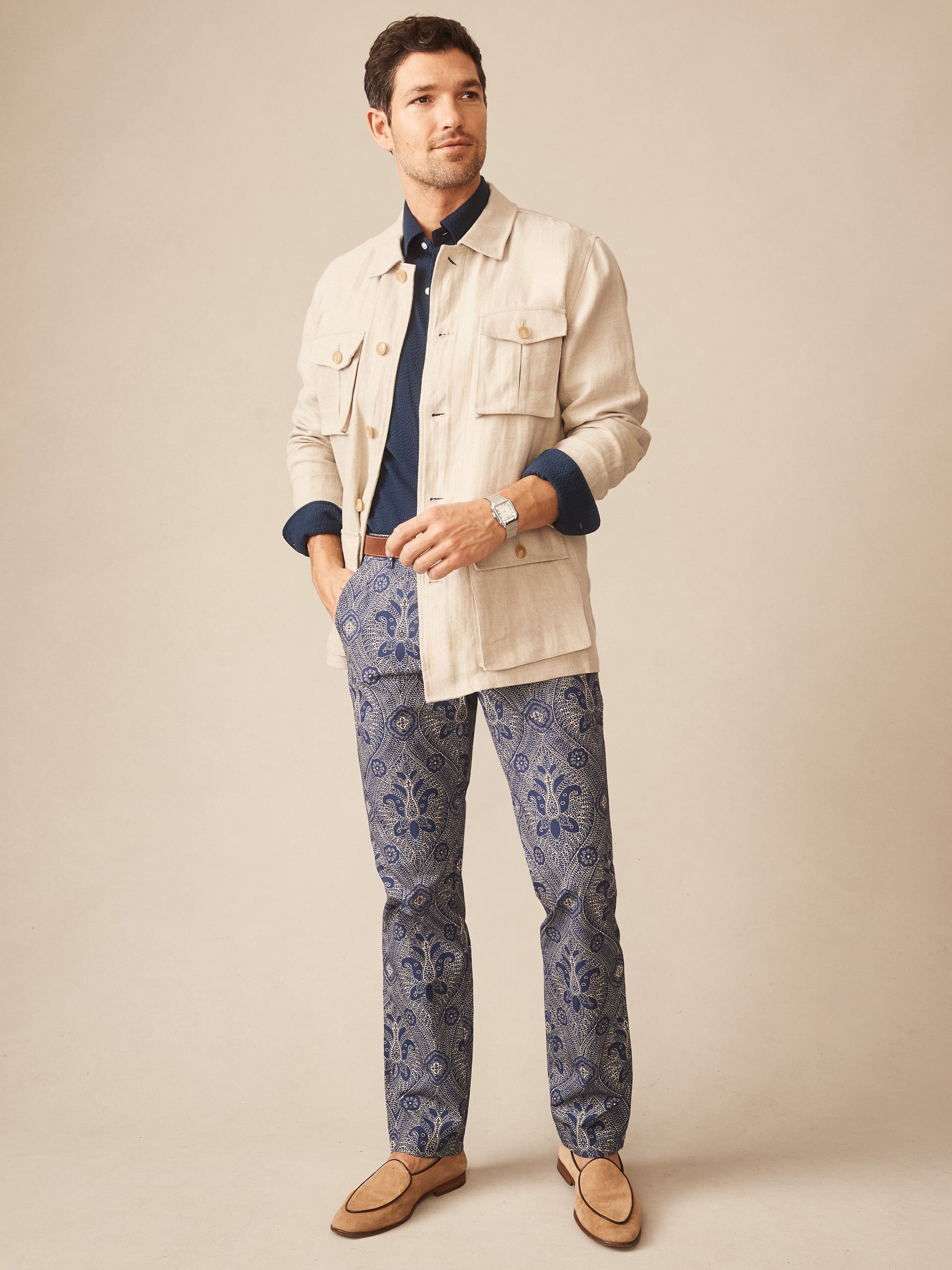 A man models the Aston Linen Safari Jacket in Herringbone by J.McLaughlin, paired with patterned pants and suede loafers, showcasing effortlessly stylish, enduring designs and innovative fabrics. A destination for defining style. Life, Well-Lived.