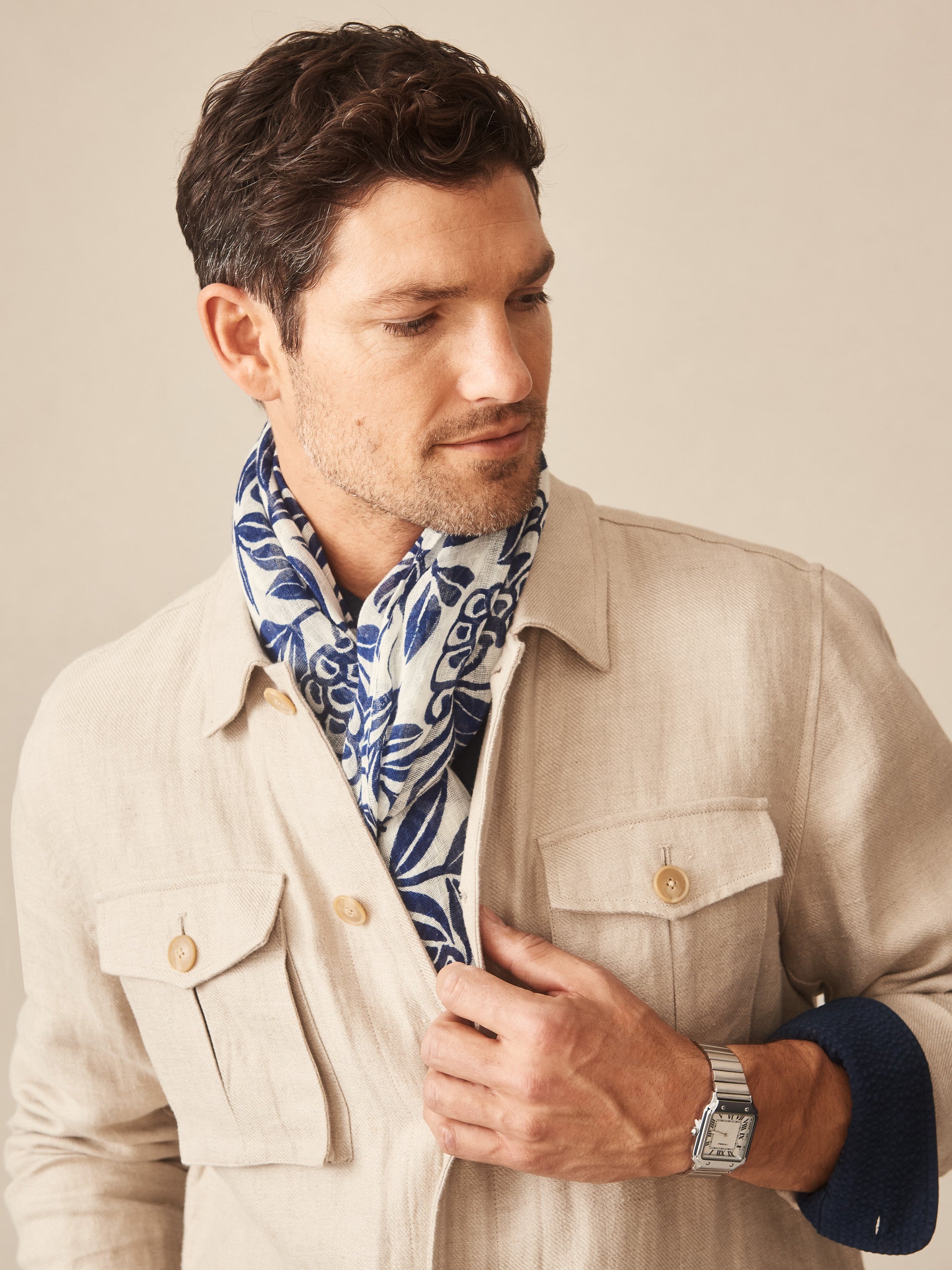 Man wearing a beige jacket and a Men's Linen Scarf in Modern Dahlia featuring blue floral patterns, showcasing J.McLaughlin’s effortlessly stylish, enduring designs with innovative fabrics.
