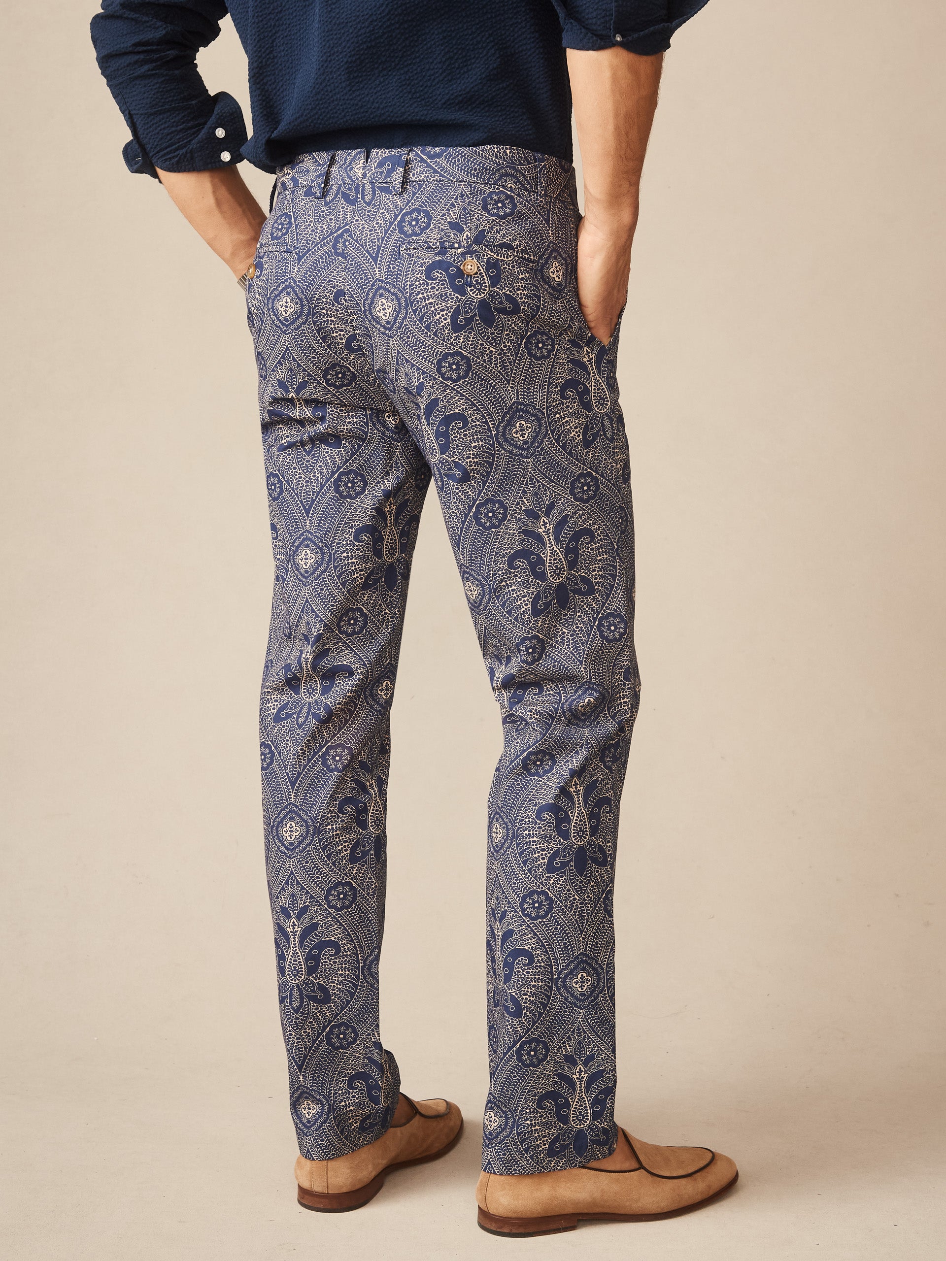 Man wearing Madison Fancy Pants in Baroque Hall by J.McLaughlin, featuring an intricate blue and beige pattern, paired with a navy shirt and tan shoes, showcasing effortlessly stylish, enduring designs and innovative fabrics. A destination for defining style. Life, Well-Lived.