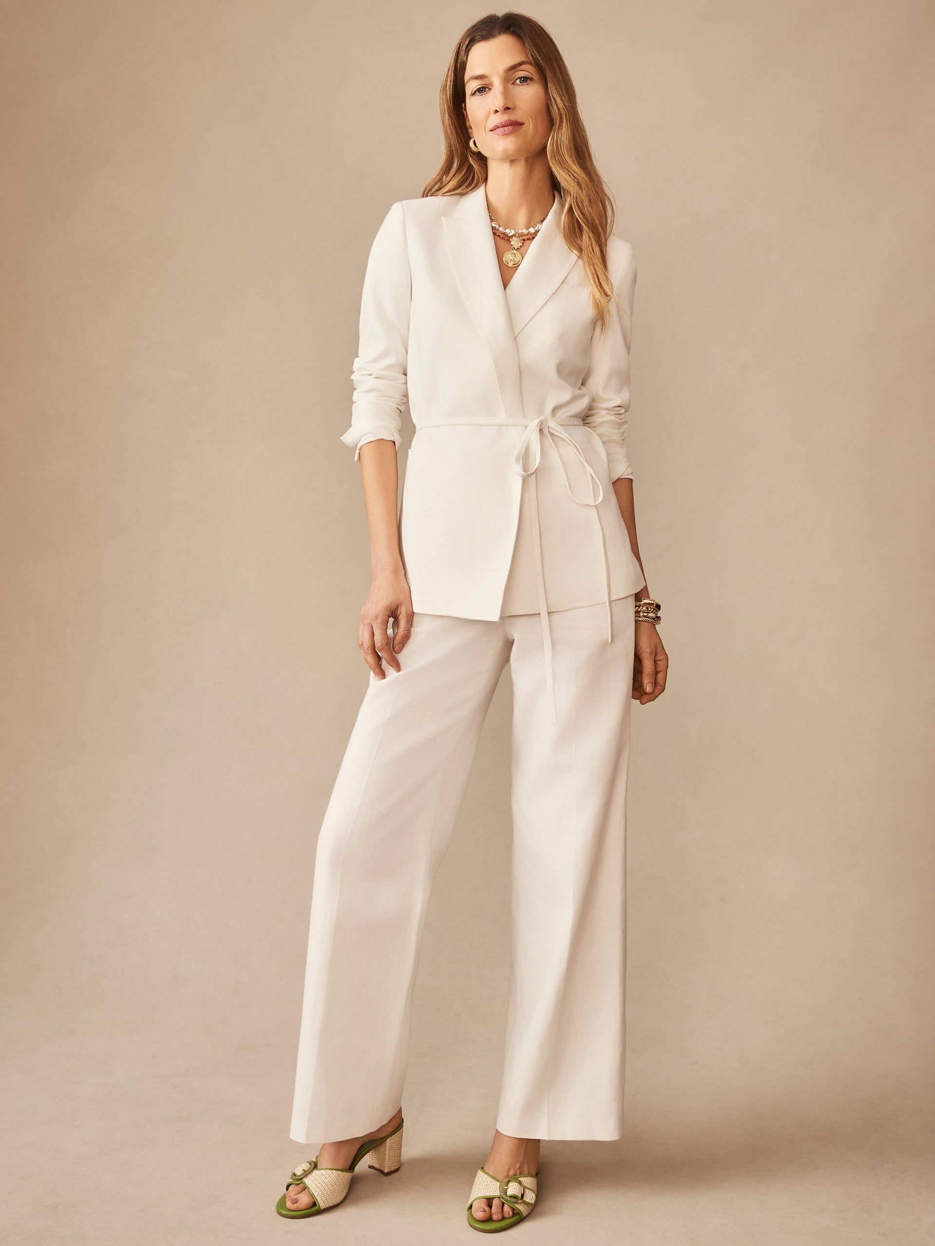 Woman wearing J.McLaughlin Blythe Linen Blend Pants in ivory paired with a matching belted blazer, styled with layered necklaces and green heeled sandals, showcasing effortlessly stylish and enduring designs.