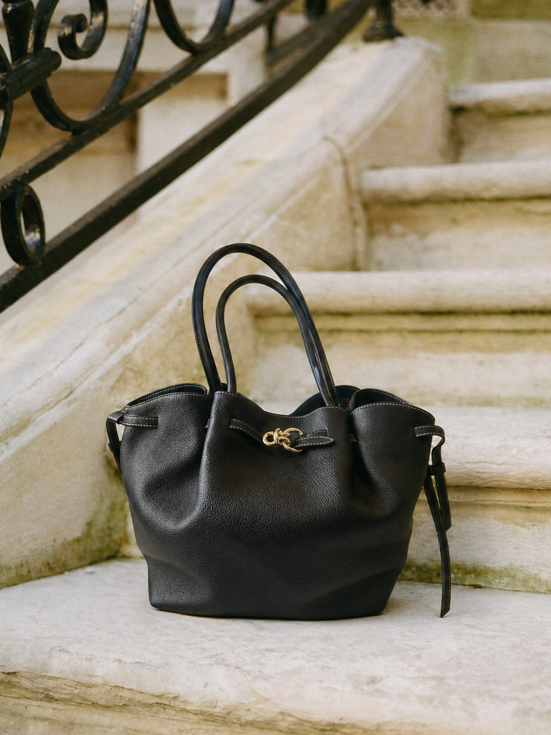 The Lex Leather Tote Bag by J.McLaughlin in black leather with a sleek design, featuring soft handles and gold-tone hardware, resting on stone steps near ornate railing.
