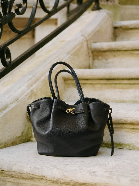 The Lex Leather Tote Bag by J.McLaughlin in black leather with a sleek design, featuring soft handles and gold-tone hardware, resting on stone steps near ornate railing.