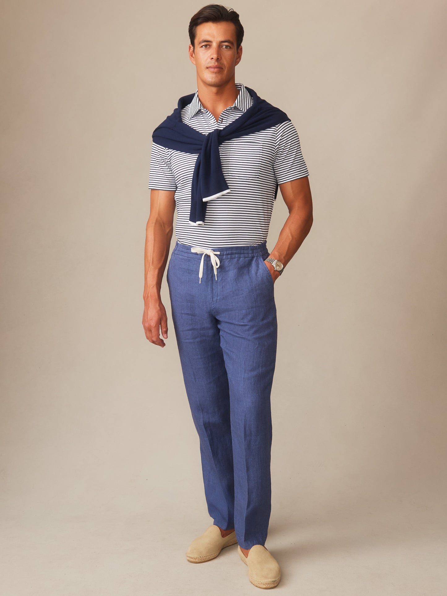 Model wearing J.McLaughlin Bleeker Linen Fancy Pants in denim blue paired with a striped polo and a navy sweater draped over shoulders, showcasing effortlessly stylish, enduring designs and innovative fabrics.