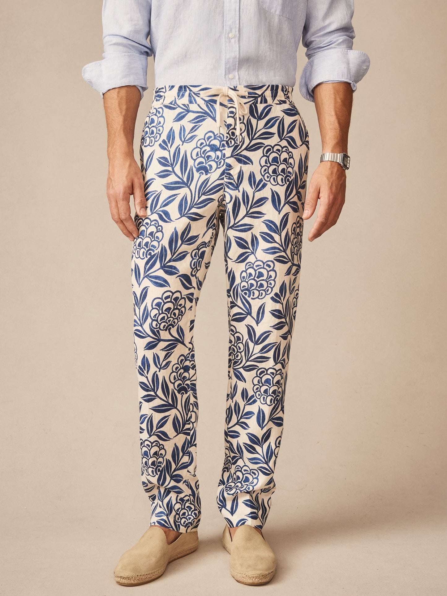 A man wearing J.McLaughlin Bleeker Linen Fancy Pants in Modern Dahlia featuring a blue botanical print on natural linen, paired with a light blue shirt and beige espadrilles, showcasing effortlessly stylish, enduring designs and innovative fabrics in a legacy of timeless fashion. A destination for defining style. Life, Well-Lived.