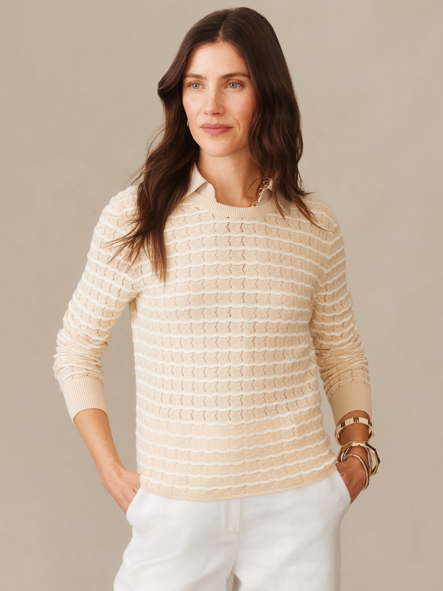 Woman wearing a beige and off-white striped Brenna Sweater in Stripe by J.McLaughlin, paired with white pants and gold jewelry, showcasing effortlessly stylish and enduring designs.