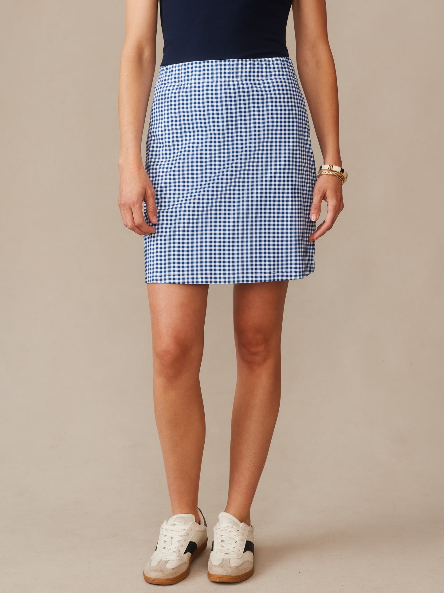 Model wearing the Briana Skort in Midi Cabana Gingham by J.McLaughlin, featuring a classic navy and white gingham pattern paired with casual white sneakers, showcasing effortlessly stylish and enduring designs in J. McLaughlin’s legacy of timeless fashion, a destination for defining style. Life, Well-Lived.