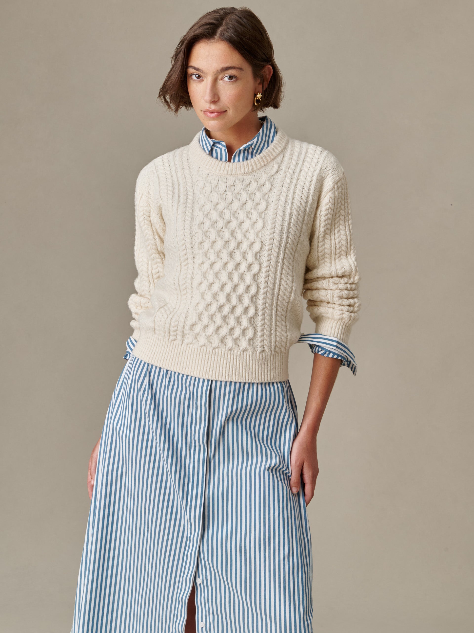 A woman wearing a cream Brigette Sweater by J.McLaughlin, featuring intricate cable knit design, styled over a blue and white striped shirt dress, reflecting enduring style and craftsmanship.