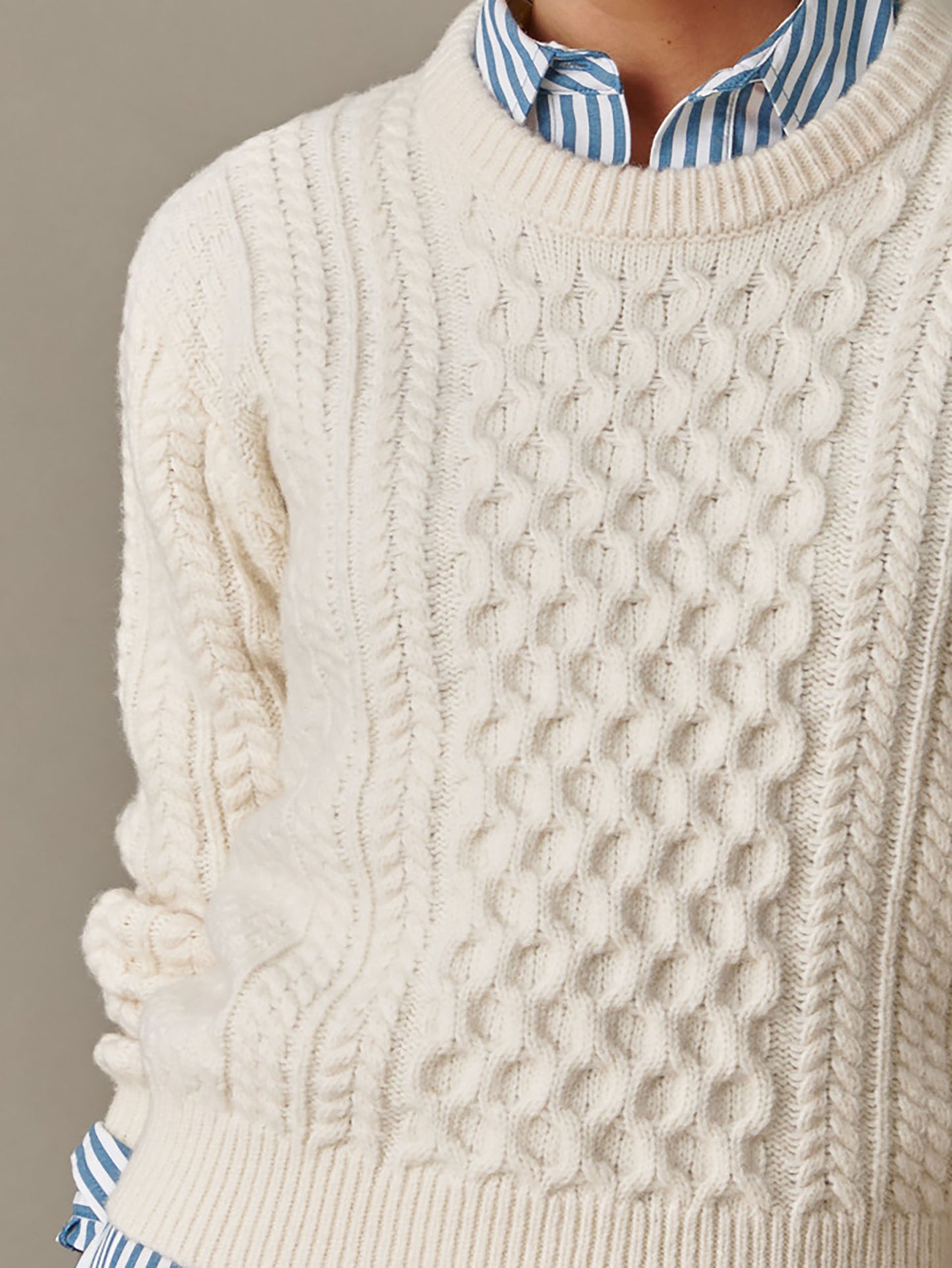 Close-up of a cozy cream cable-knit Brigette Sweater by J.McLaughlin, layered over a blue and white striped shirt, showcasing timeless style and enduring craftsmanship.
