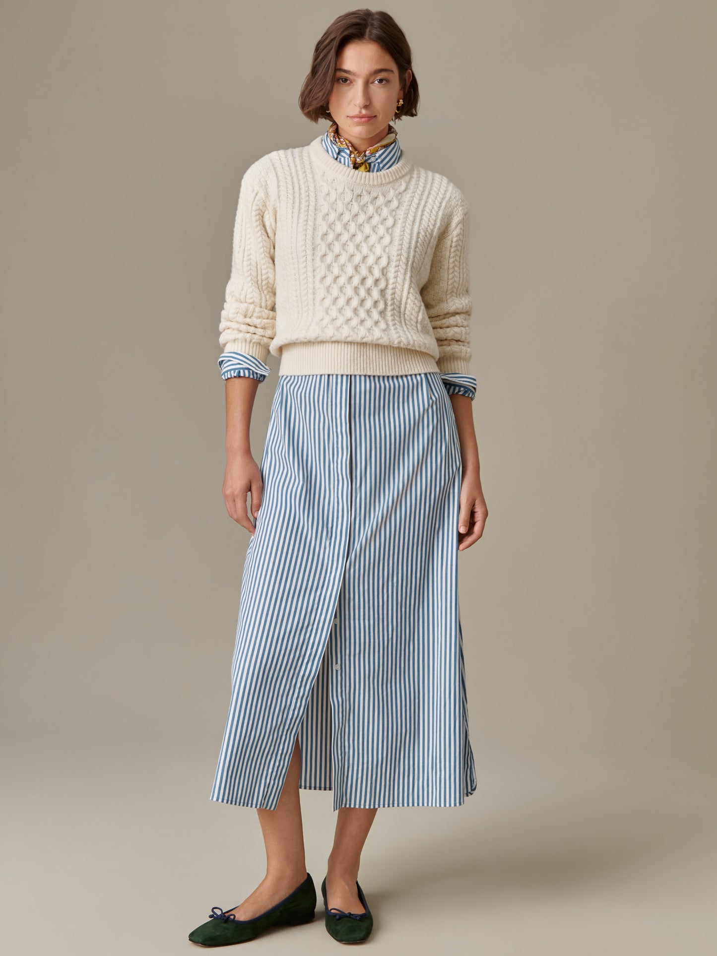 Woman wearing a cream Brigette Sweater by J.McLaughlin with cable knit detail, layered over a striped collared shirt and paired with a blue and white striped midi skirt and dark ballet flats.