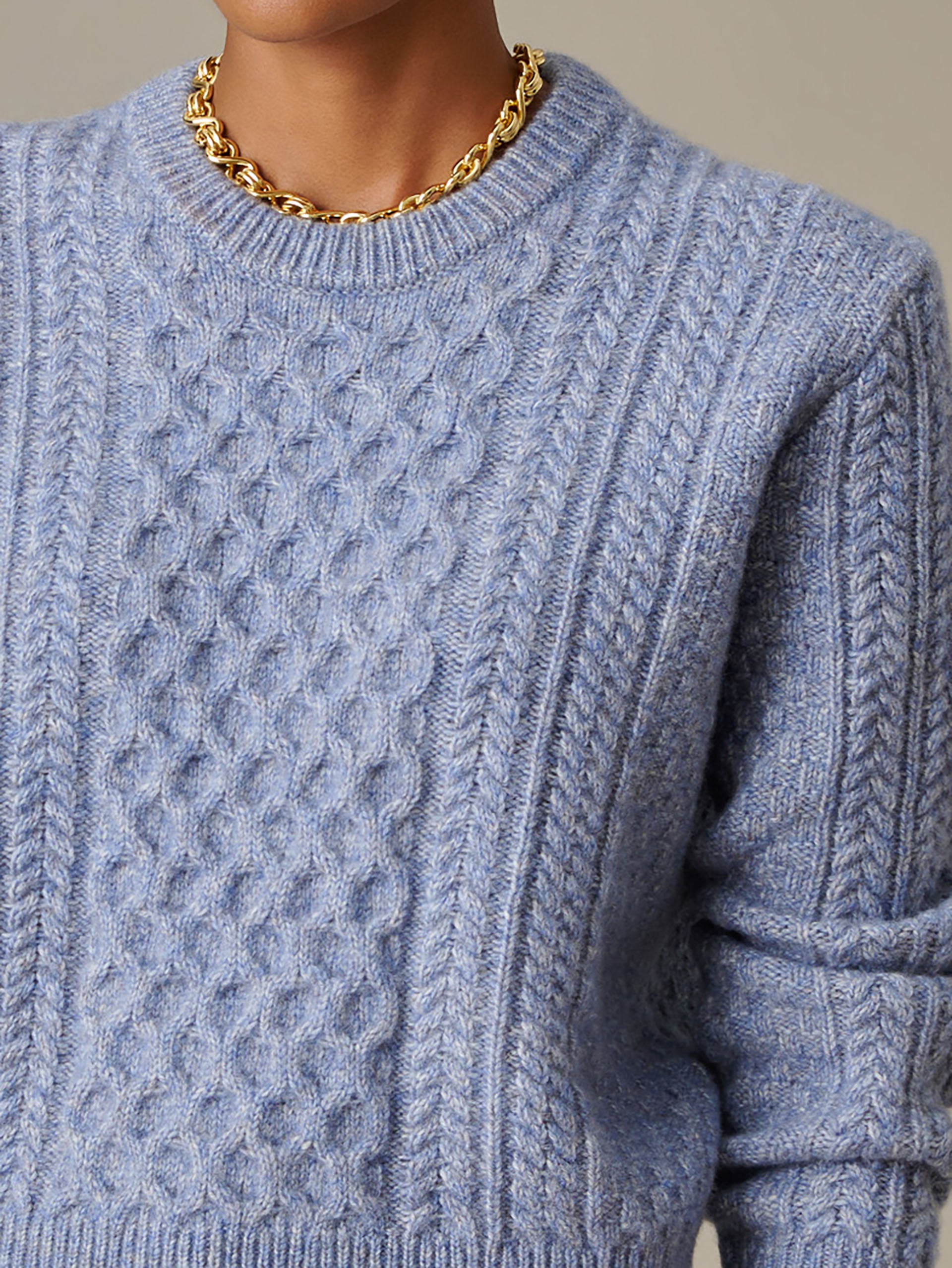 Close-up of a person wearing a light blue Brigette Sweater by J.McLaughlin, featuring intricate cable knit patterns and a cozy, timeless design paired with a gold chain necklace.