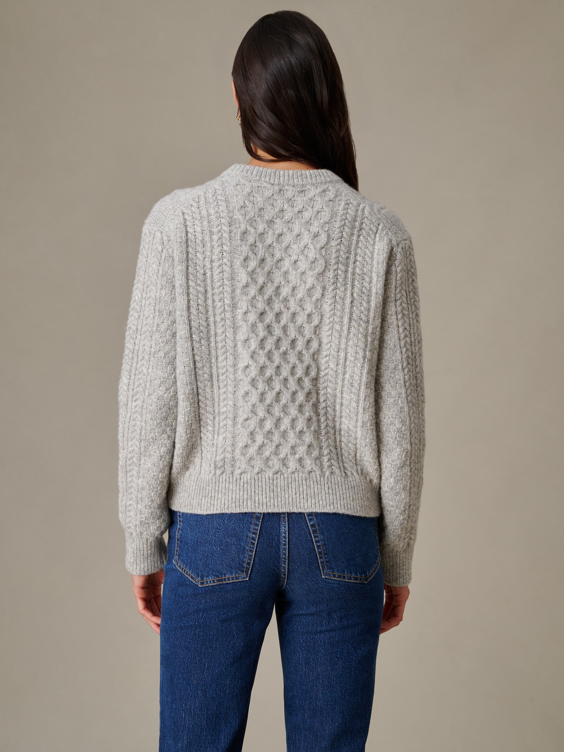 Woman wearing a light gray Brigette Sweater by J.McLaughlin featuring intricate cable knit design, paired with classic blue jeans, showcasing enduring style and craftsmanship.