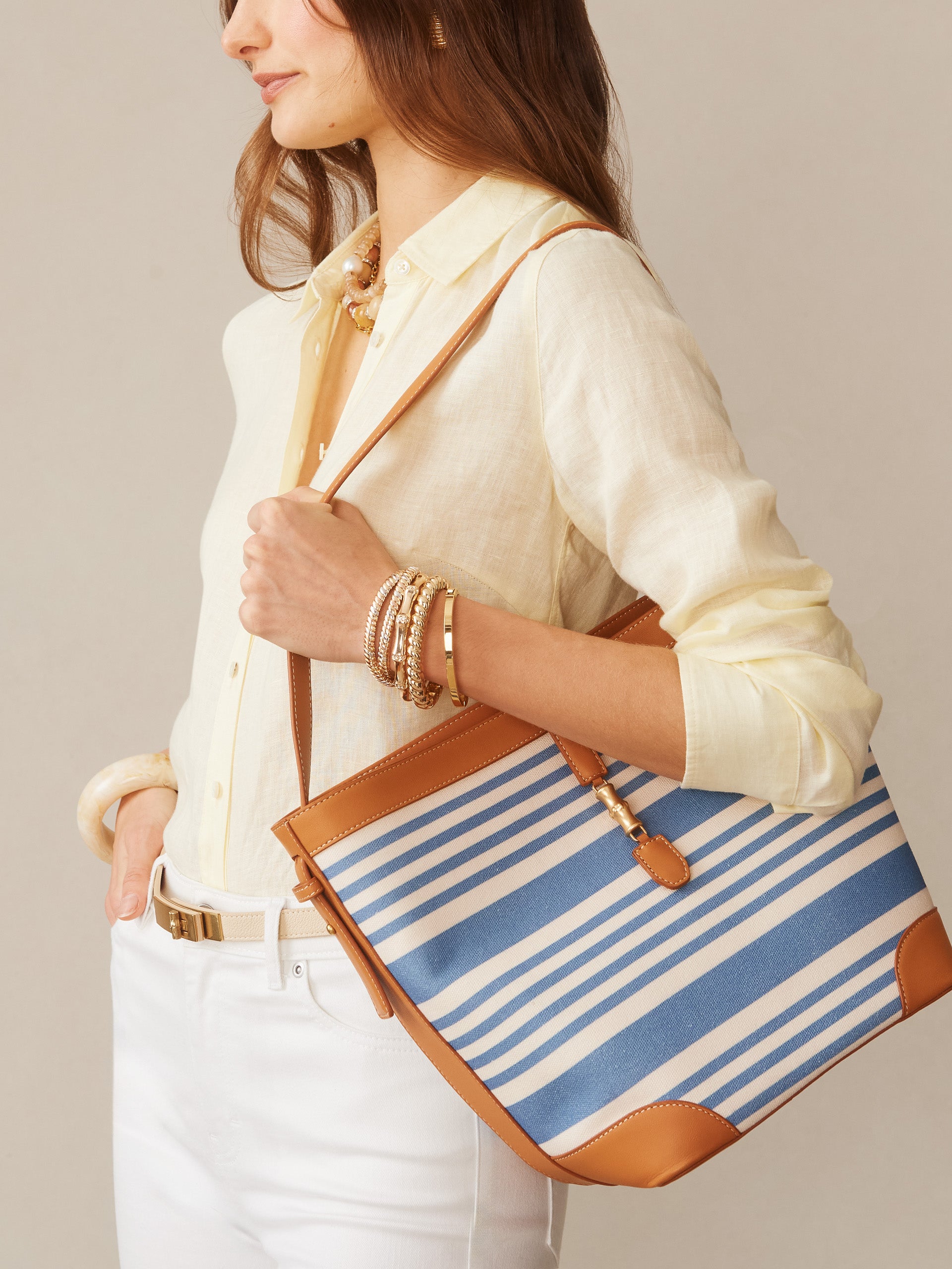 Woman wearing a light yellow blouse and white pants carrying the Crawford Handbag in Stripe by J.McLaughlin with blue and white stripes and tan leather accents, showcasing effortlessly stylish, enduring designs.