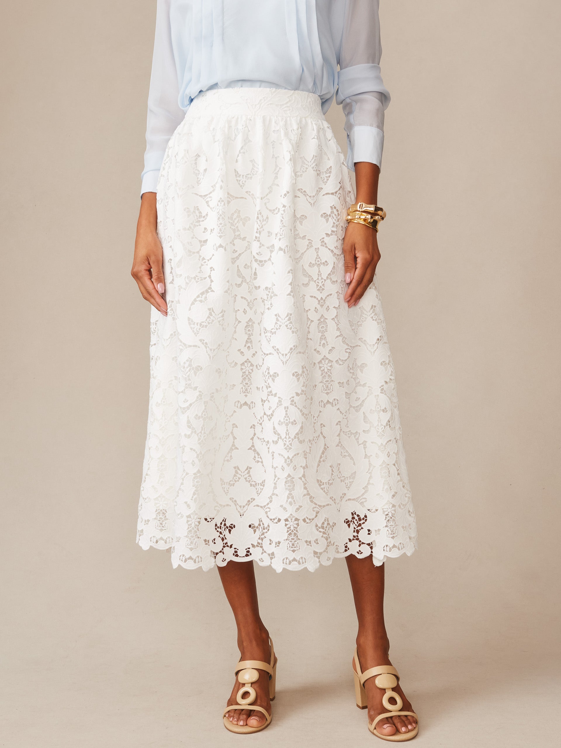 A model wearing the Callie Lace Skirt by J.McLaughlin in off-white, styled with a light blue blouse and beige sandals, showcasing effortlessly stylish, enduring designs and a legacy of timeless fashion.