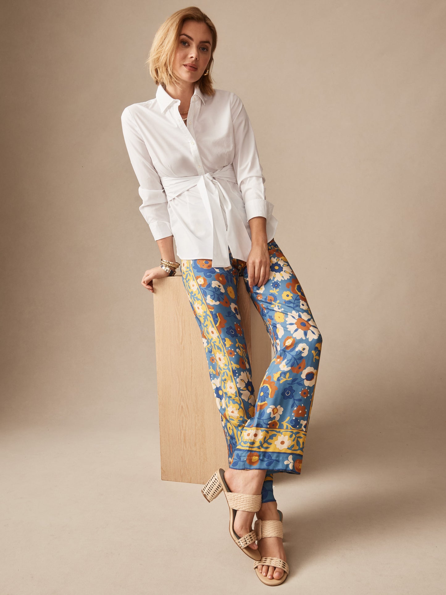 Woman wearing J.McLaughlin Carter Silk Blend Pants in Floral Daydream Edge with blue, yellow, and brown floral patterns paired with a classic white blouse and woven heeled sandals, showcasing effortlessly stylish, enduring designs crafted from innovative fabrics.