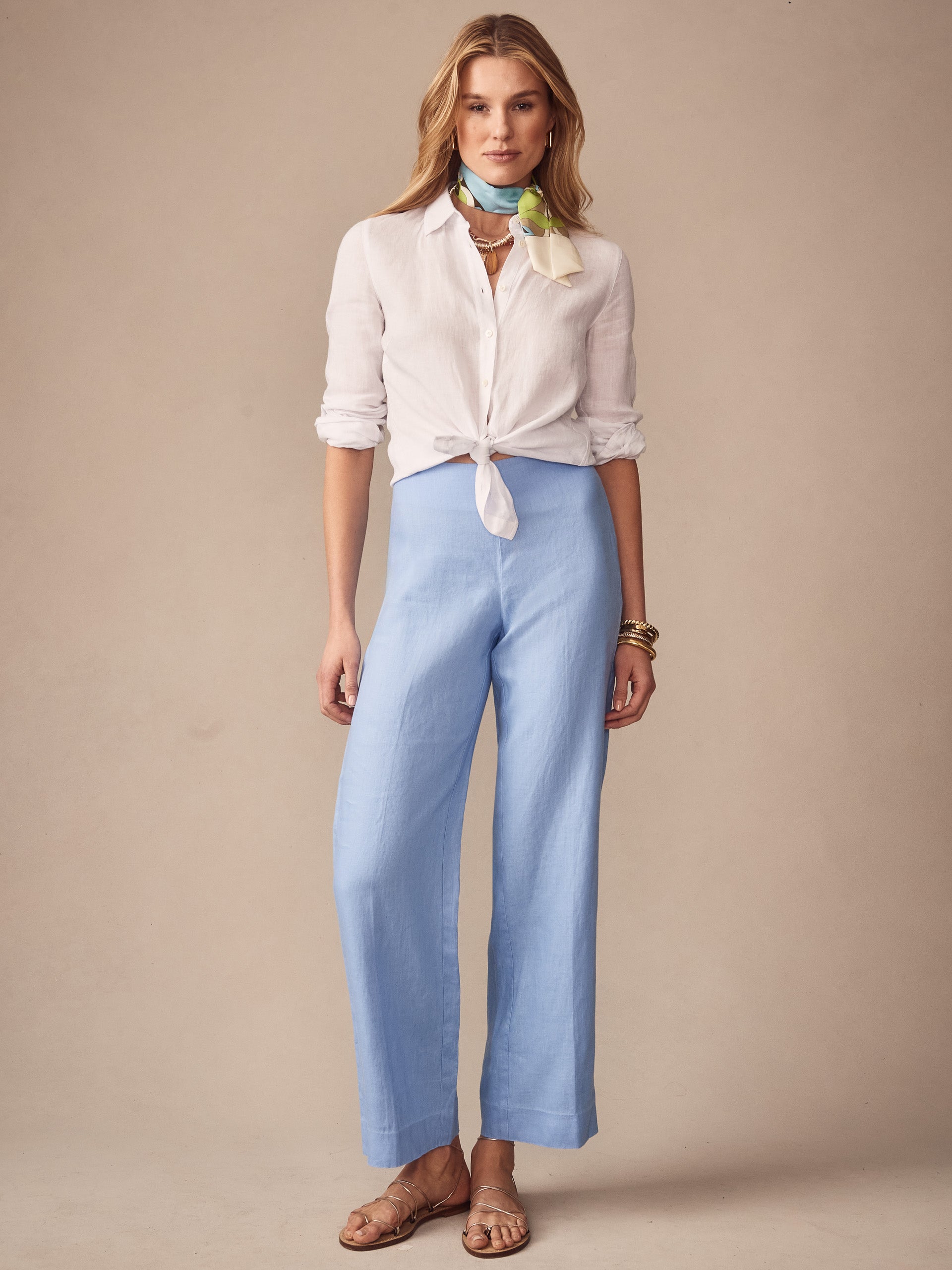 A woman wears J.McLaughlin Carter Linen Pants in soft blue paired with a tied white blouse, scarf, and sandals, showcasing effortlessly stylish, enduring designs for a life well-lived.