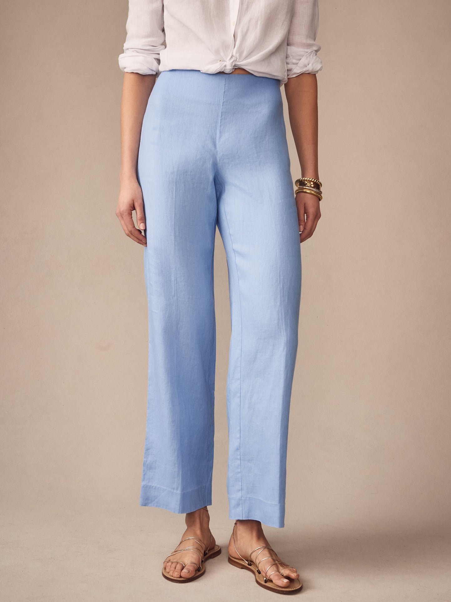 A model wearing J.McLaughlin Carter Linen Pants in a soft cornflower blue, paired with a rolled-sleeve white shirt and gold bracelets, showcasing effortlessly stylish, enduring designs with innovative fabrics.