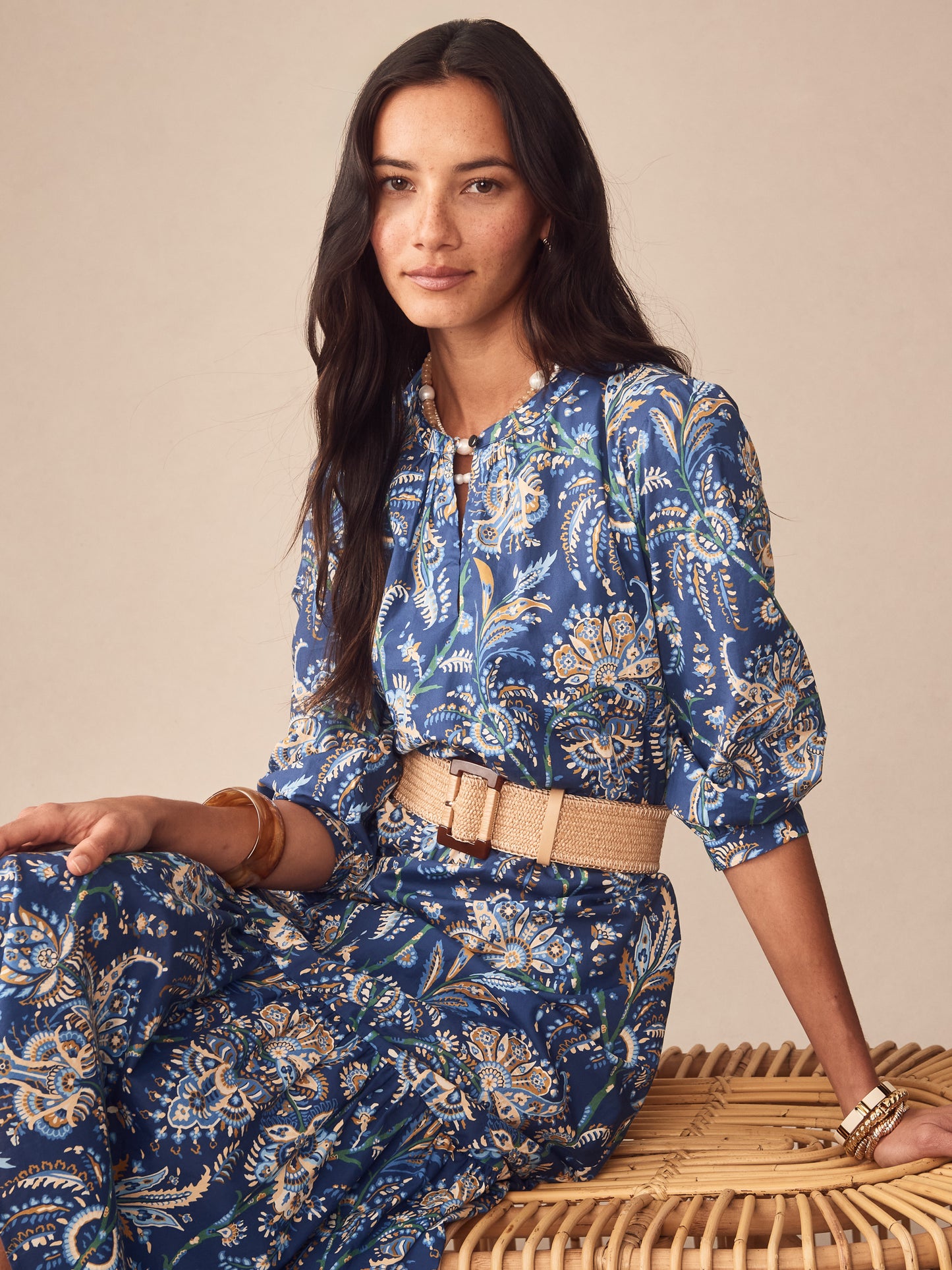 Woman seated wearing the Cerina Dress in Maxi Heirloom Paisley by J.McLaughlin, featuring a navy floral pattern paired with a woven belt, embodying effortlessly stylish and enduring designs.