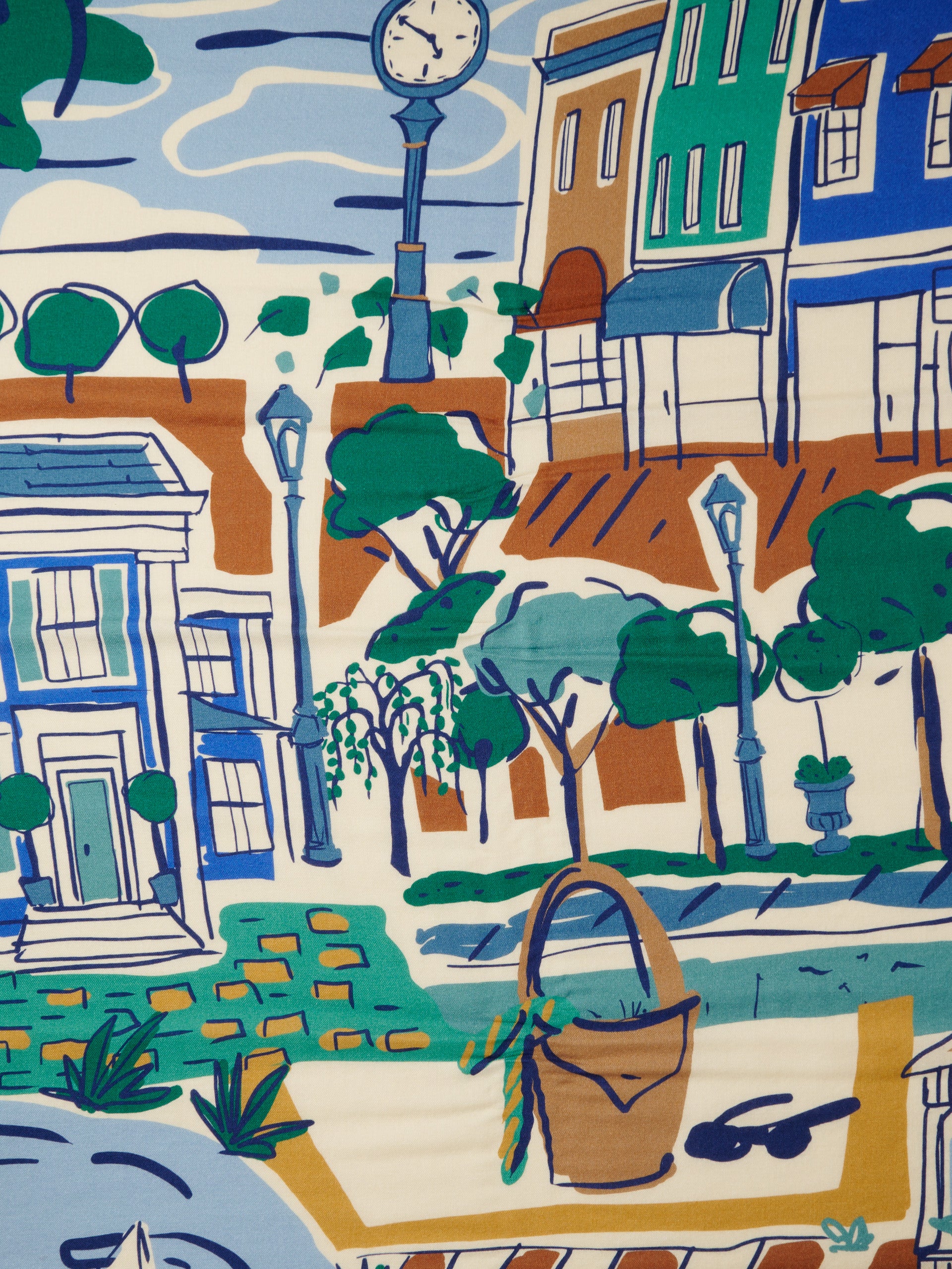 The Charlotte Scarf in Village Stroll Border by J.McLaughlin featuring a colorful illustrated village street scene with trees buildings and a bicycle basket