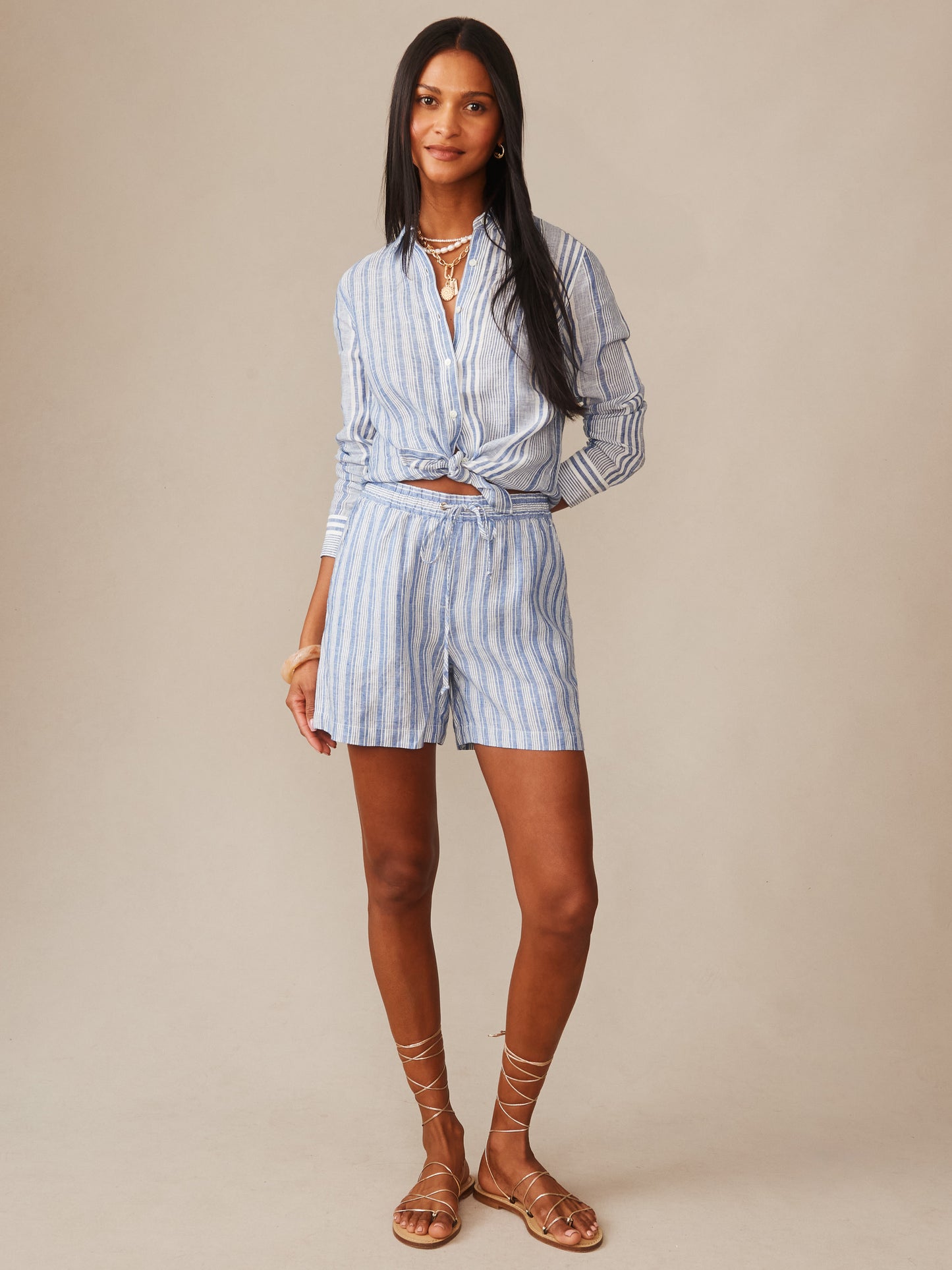Woman wearing J.McLaughlin Charlotte Linen Shorts in Stripe paired with a matching striped linen shirt, styled with layered necklaces and sandals, showcasing effortlessly stylish, enduring designs.