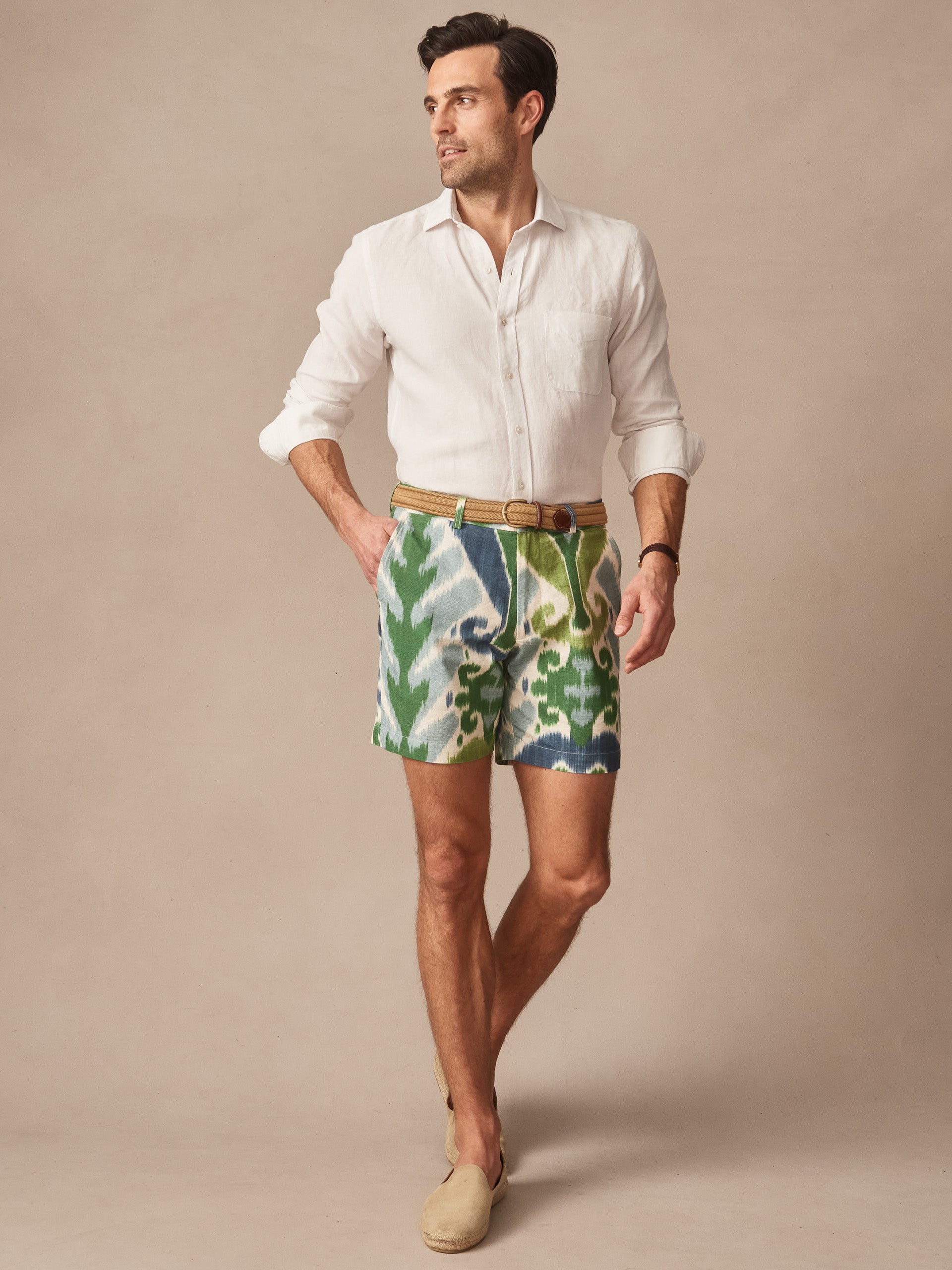 A man modeling J.McLaughlin's Chatham Shorts in Ikat featuring green and blue patterns, paired with a white shirt and beige belt, showcasing effortlessly stylish shorts for enduring designs.